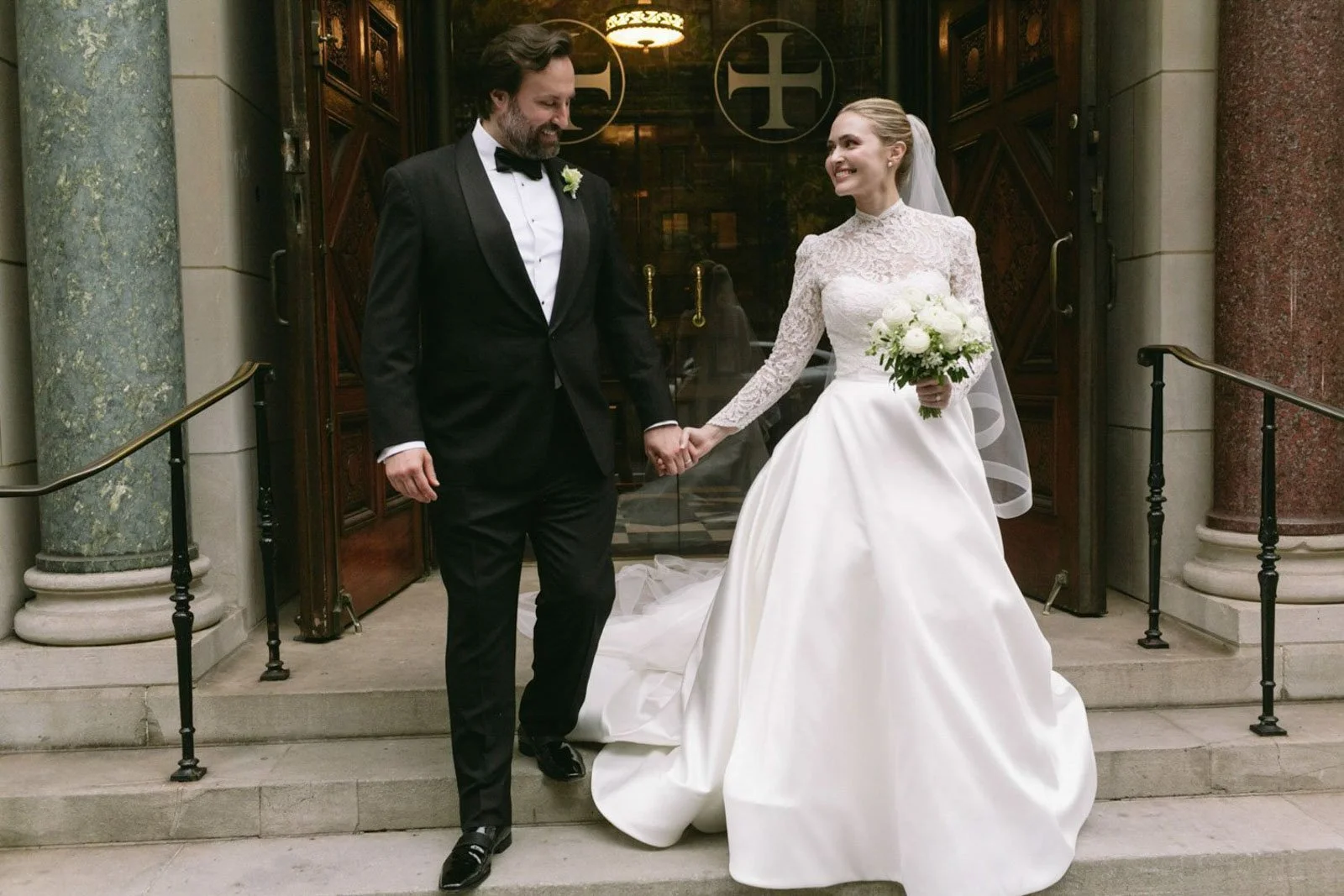 A newlywed couple holding hands in front of a church entrance. The groom is wearing a black tuxedo with a bow tie and the bride in a white lace long-sleeve wedding gown holding a bouquet of white flowers. They are smiling at each other.