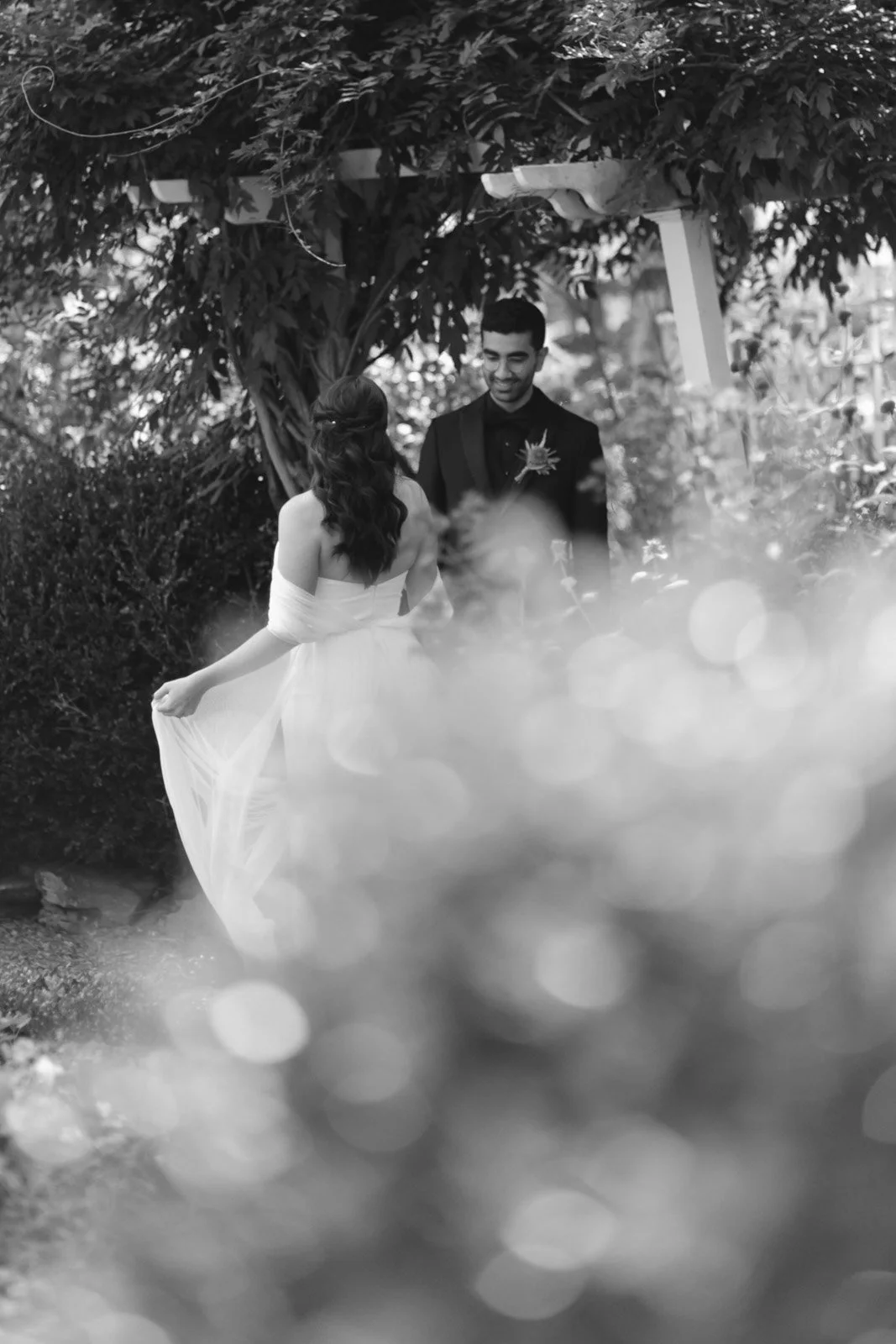 A black-and-white photo of a bride and groom exchanging vows outdoors under a wooden arch, surrounded by lush greenery, with the bride lifting her dress and the groom smiling.