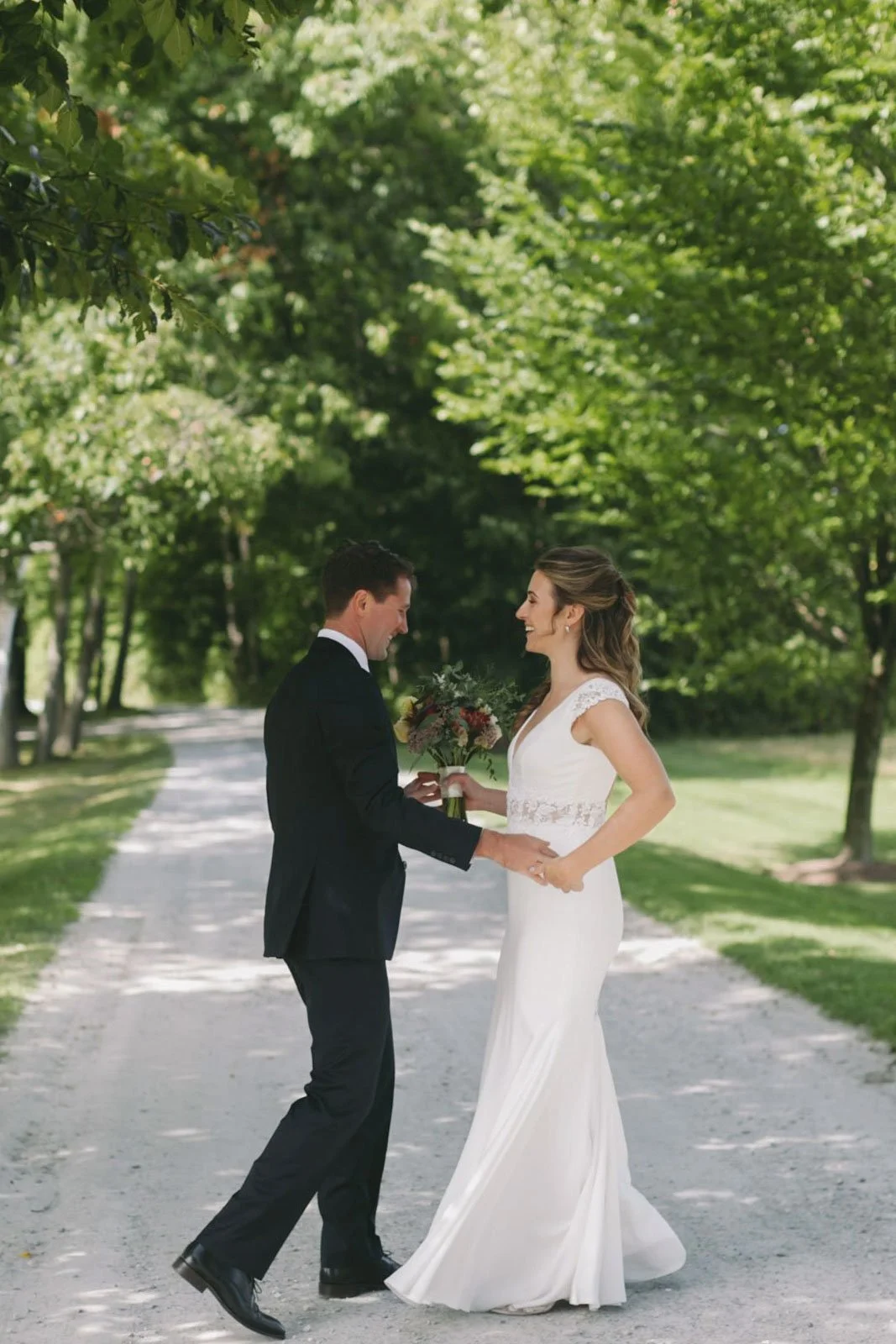 A bride and groom in wedding attire sharing a moment outdoors on a tree-lined path, with the groom holding a bouquet and both smiling.