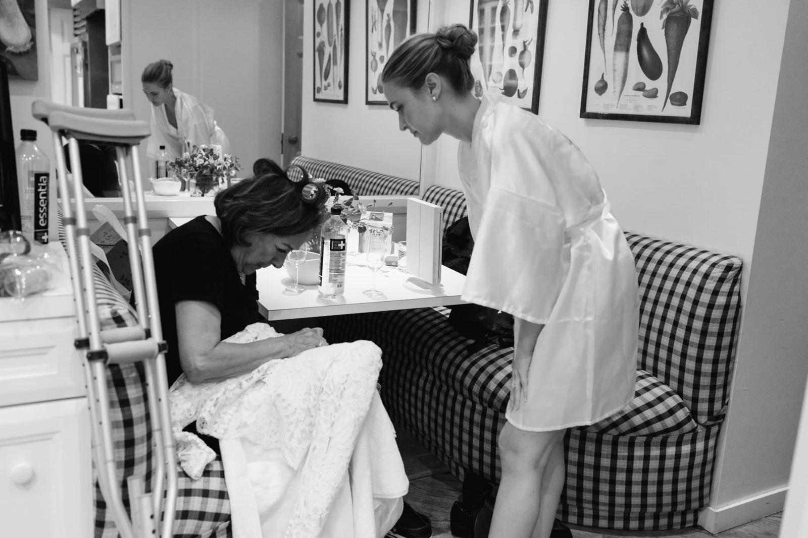 A woman in a white robe is leaning over a seated woman in a black dress, who is in a hospital bed, surrounded by hospital supplies and decorations, with another woman in a white robe in the background.