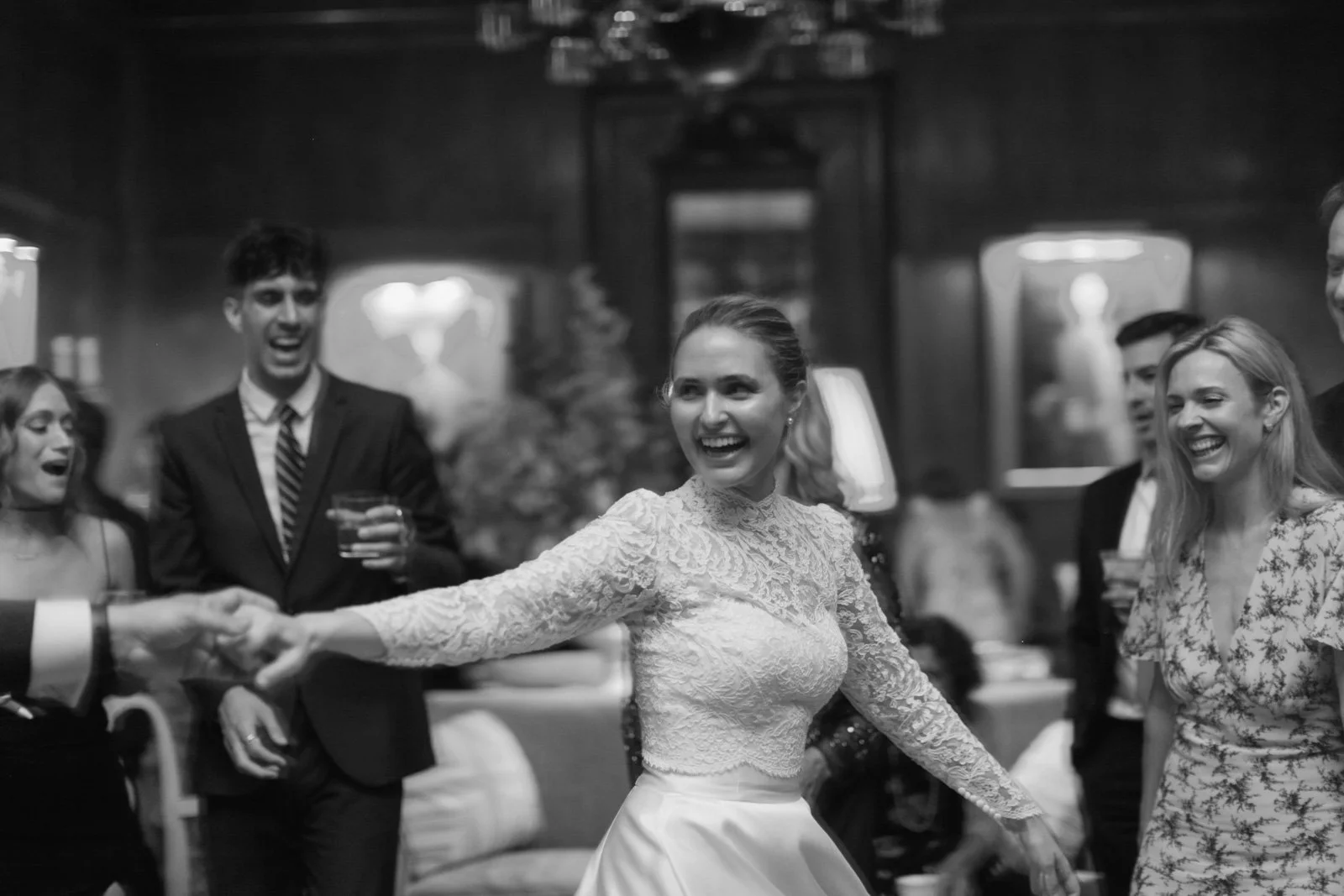 People enjoying a dance at a celebration or event, with a woman in a lace top and satin skirt dancing surrounded by smiling friends in formal attire.
