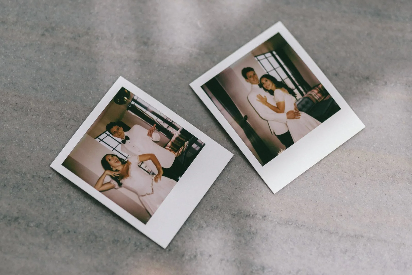 Two instant photographs on a gray surface; one features a woman in a white dress with dark hair and the other a man and woman dressed in wedding attire standing by a window.