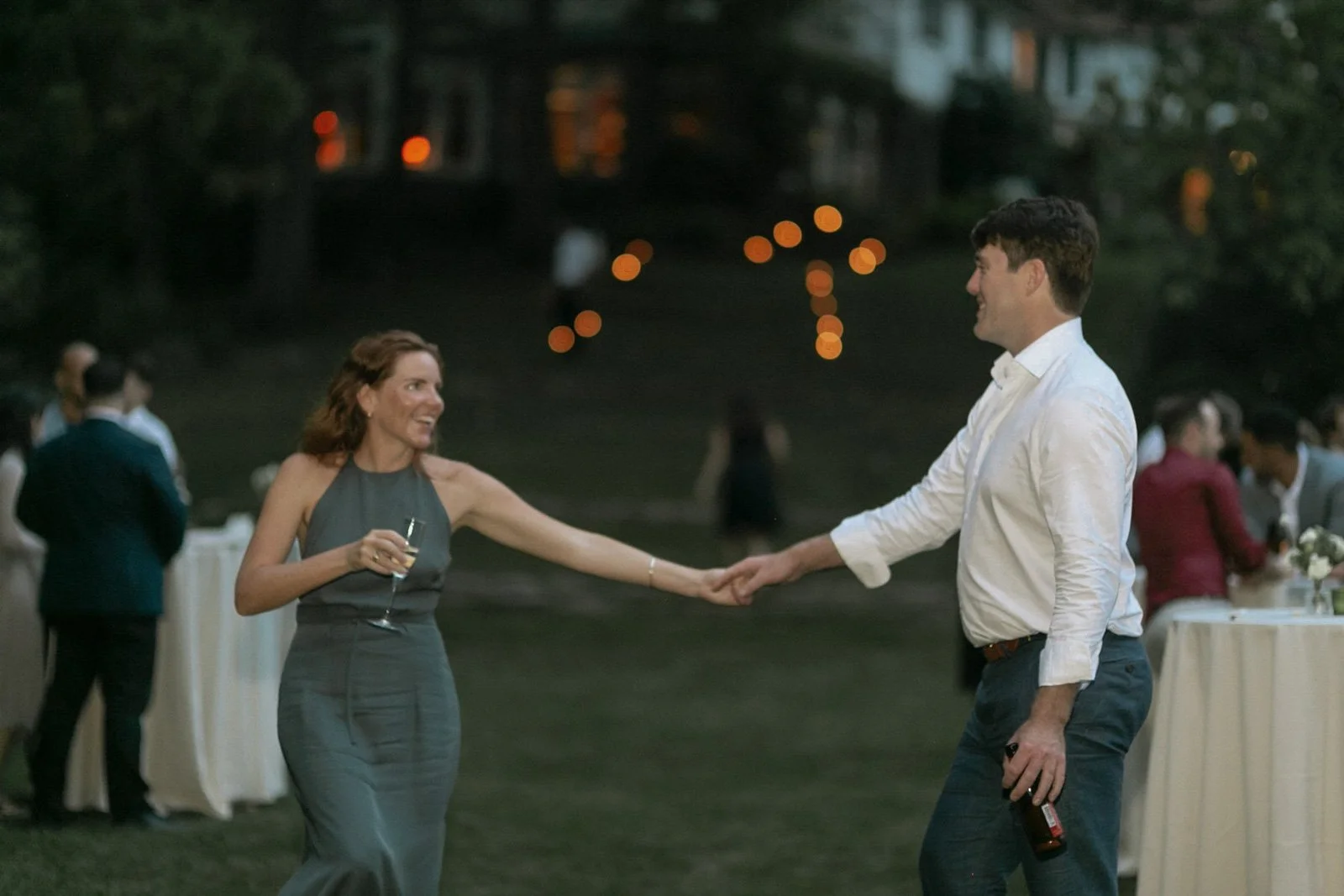A woman and a man are dancing and holding hands at an outdoor evening event. The woman is wearing a long, sleeveless gray dress and holding a glass. The man is dressed in a white shirt and dark pants, holding a bottle in his right hand. There are oth