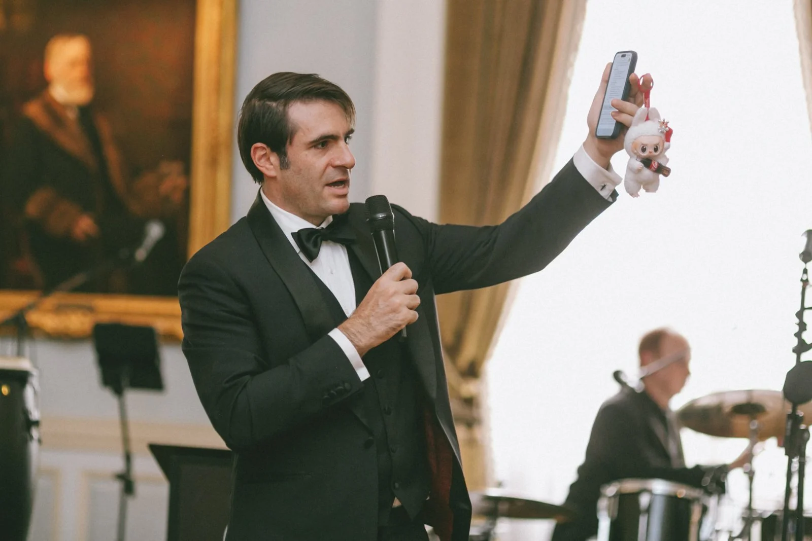 Man in tuxedo holding microphone and taking a selfie with a plush toy at a formal event with musicians and a large portrait in the background.