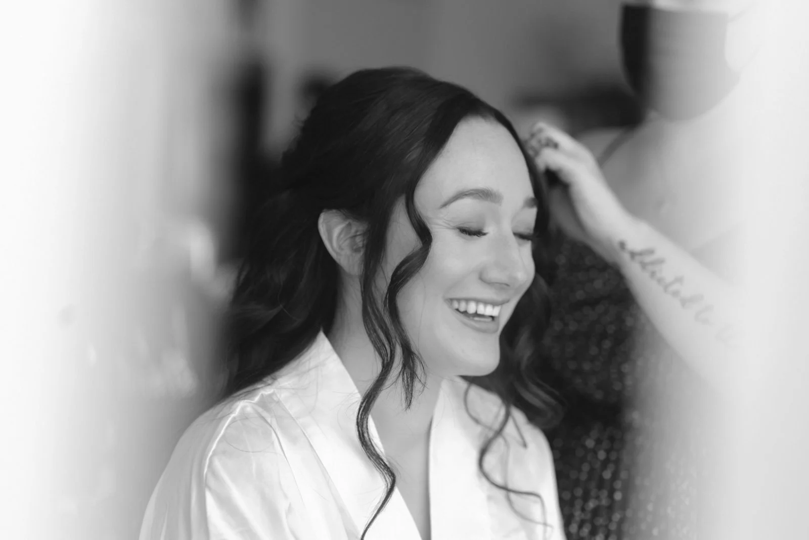 A woman with long dark hair smiling with eyes closed, having her hair styled by a person in the background.