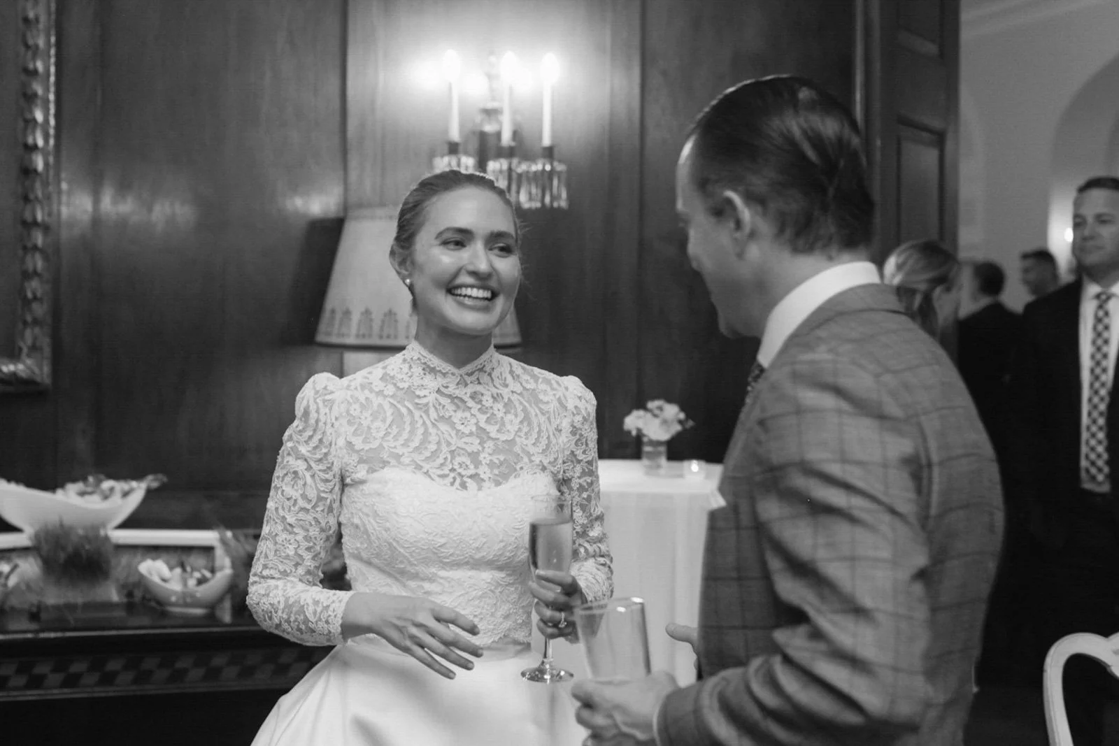 A woman in a lace wedding dress smiling and holding a glass, talking to a man in a suit at a wedding reception.