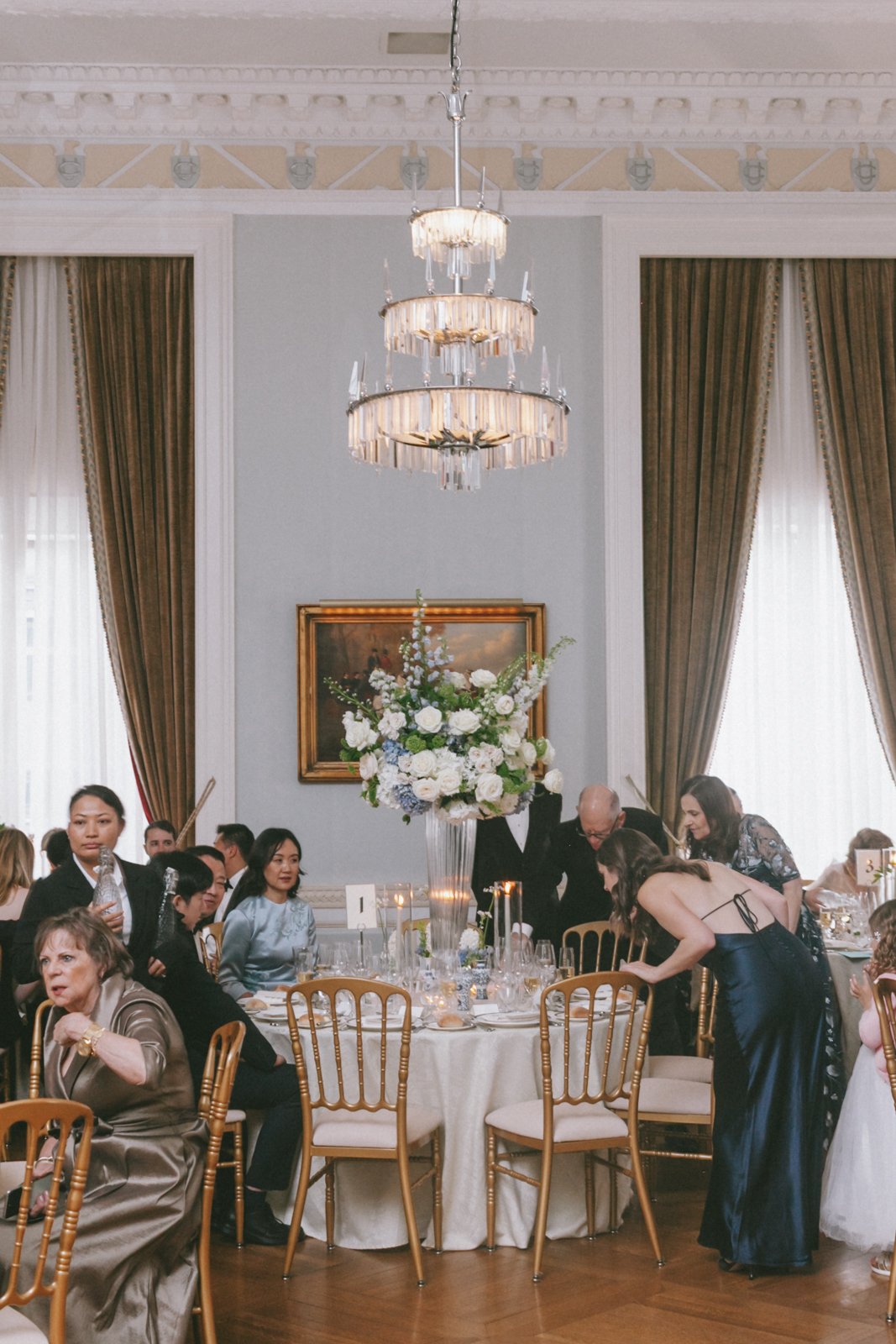 Elegant dining room with a large floral centerpiece, chandelier, and gold chairs, hosting people in formal attire seated around a round table during a special event or celebration.