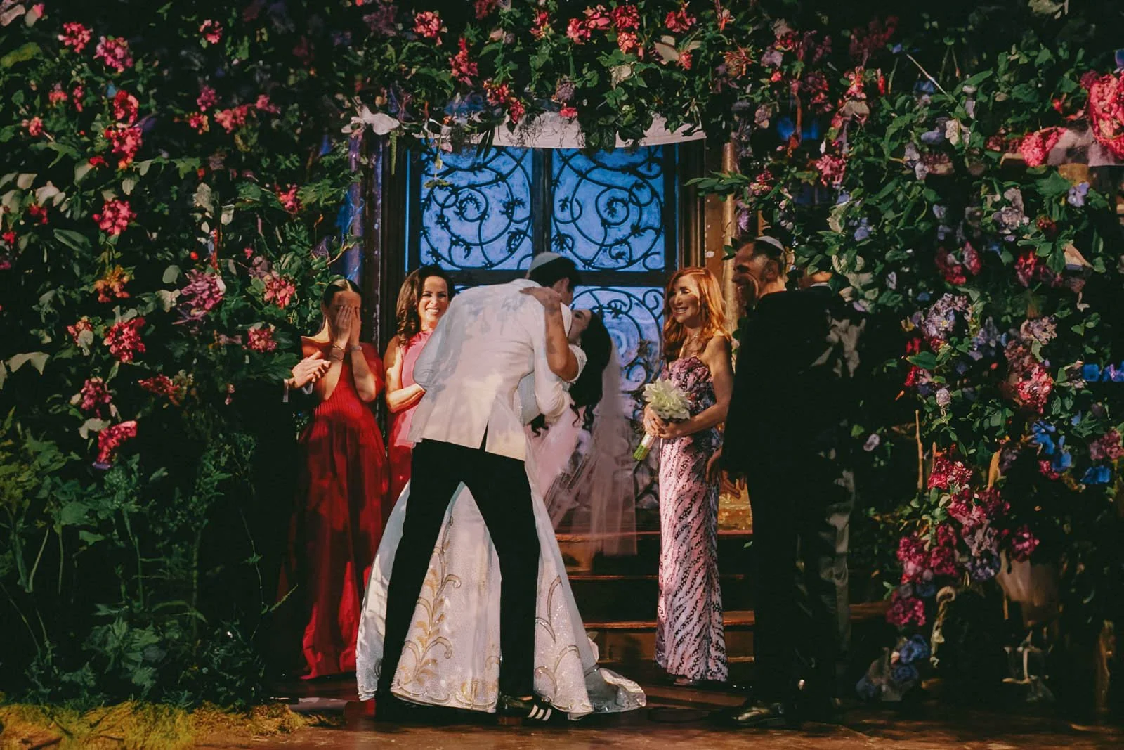 A wedding ceremony with the bride and groom kissing, surrounded by bridesmaids and groomsmen standing in front of a decorated floral backdrop.