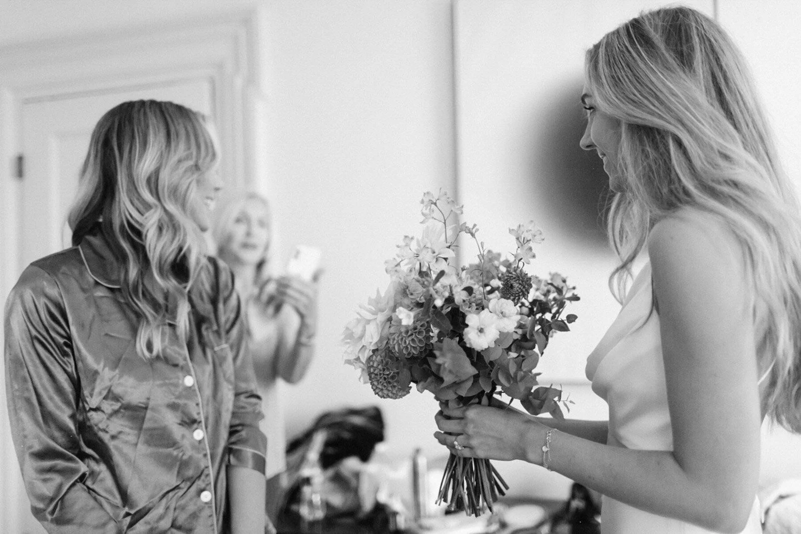 A woman with long, wavy hair is holding a bouquet of flowers and smiling, while another woman in satin pajamas is smiling and looking at her. In the background, a third woman is looking at her phone.