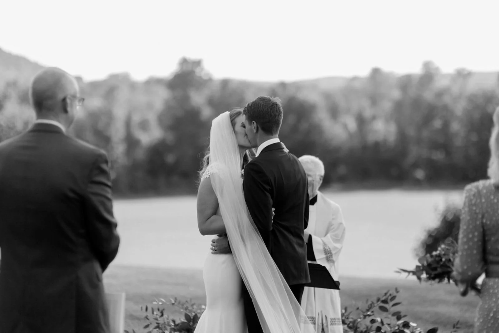 A couple kissing during their wedding ceremony outdoors with officiant and guests visible, scenic landscape background.