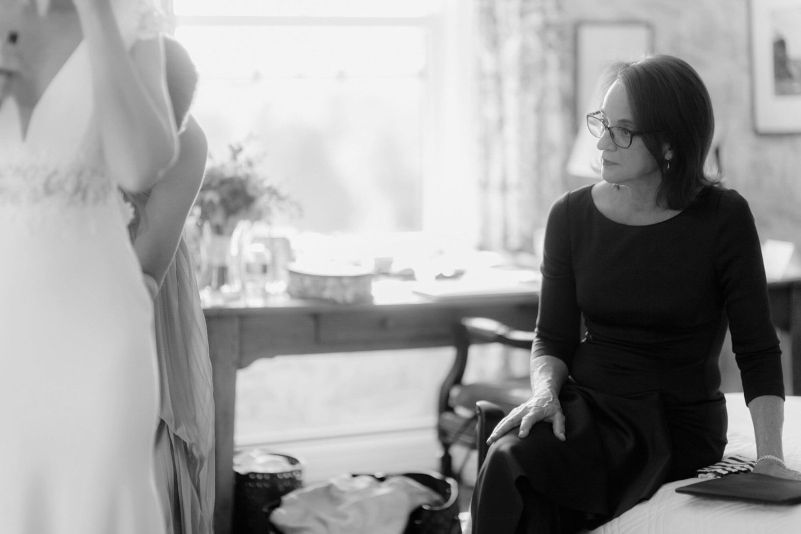 A woman with glasses sitting on a bed, looking at another person who is standing and dressed in light-colored clothing, in a room with a bright window and some furniture.