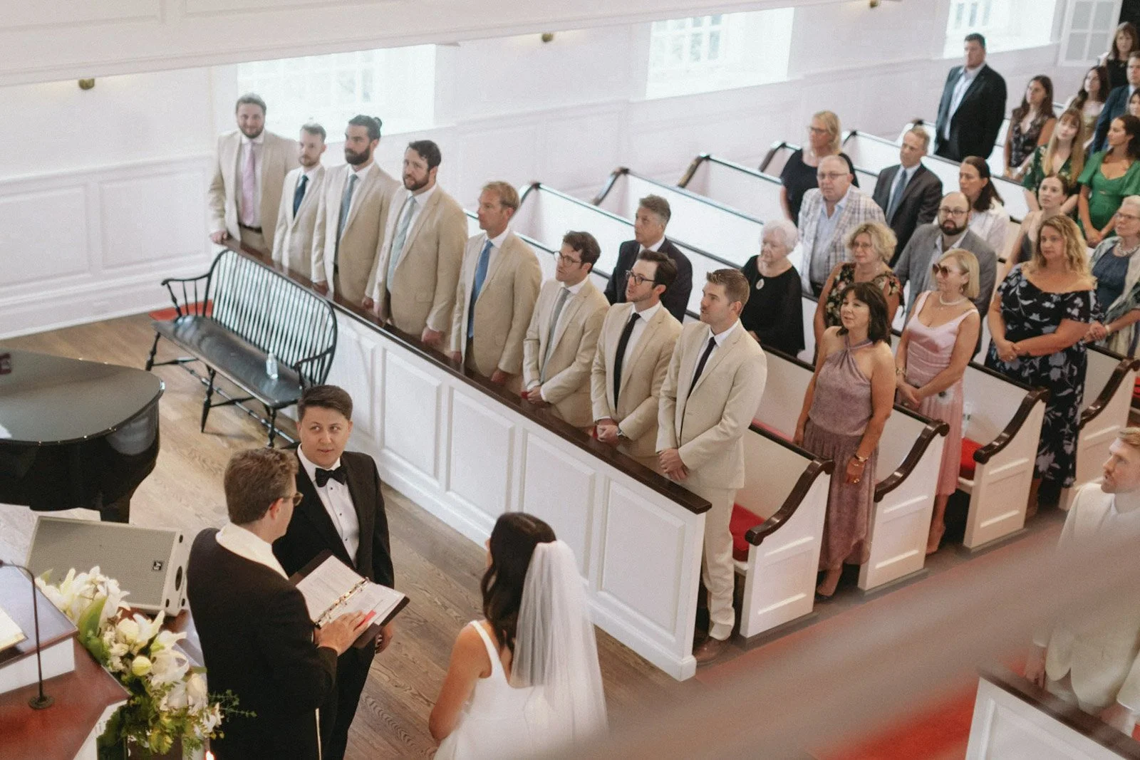 A wedding ceremony inside a church with guests standing in pews, a bride and groom at the altar, and the officiant reading from a book.