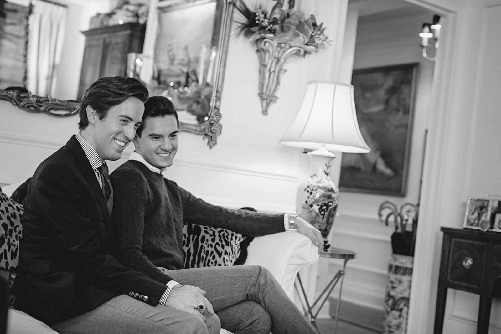 Two men in formal attire sitting on a sofa in a living room, smiling, with decorative elements including a lamp, mirror, and paintings in the background.