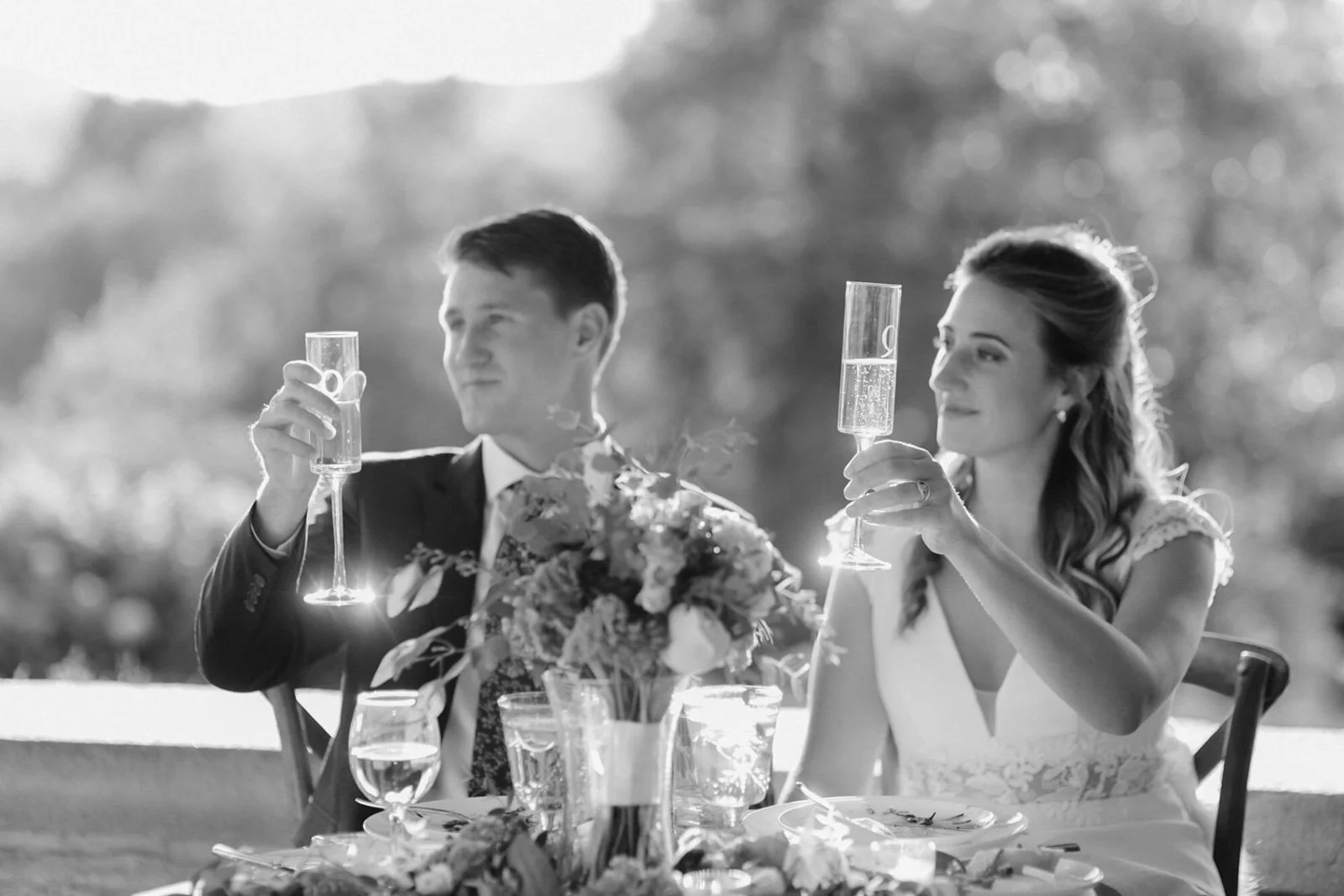 A black and white photo of a bride and groom raising glasses in toast at their wedding reception, seated at a table with floral centerpiece and table setting outdoors.