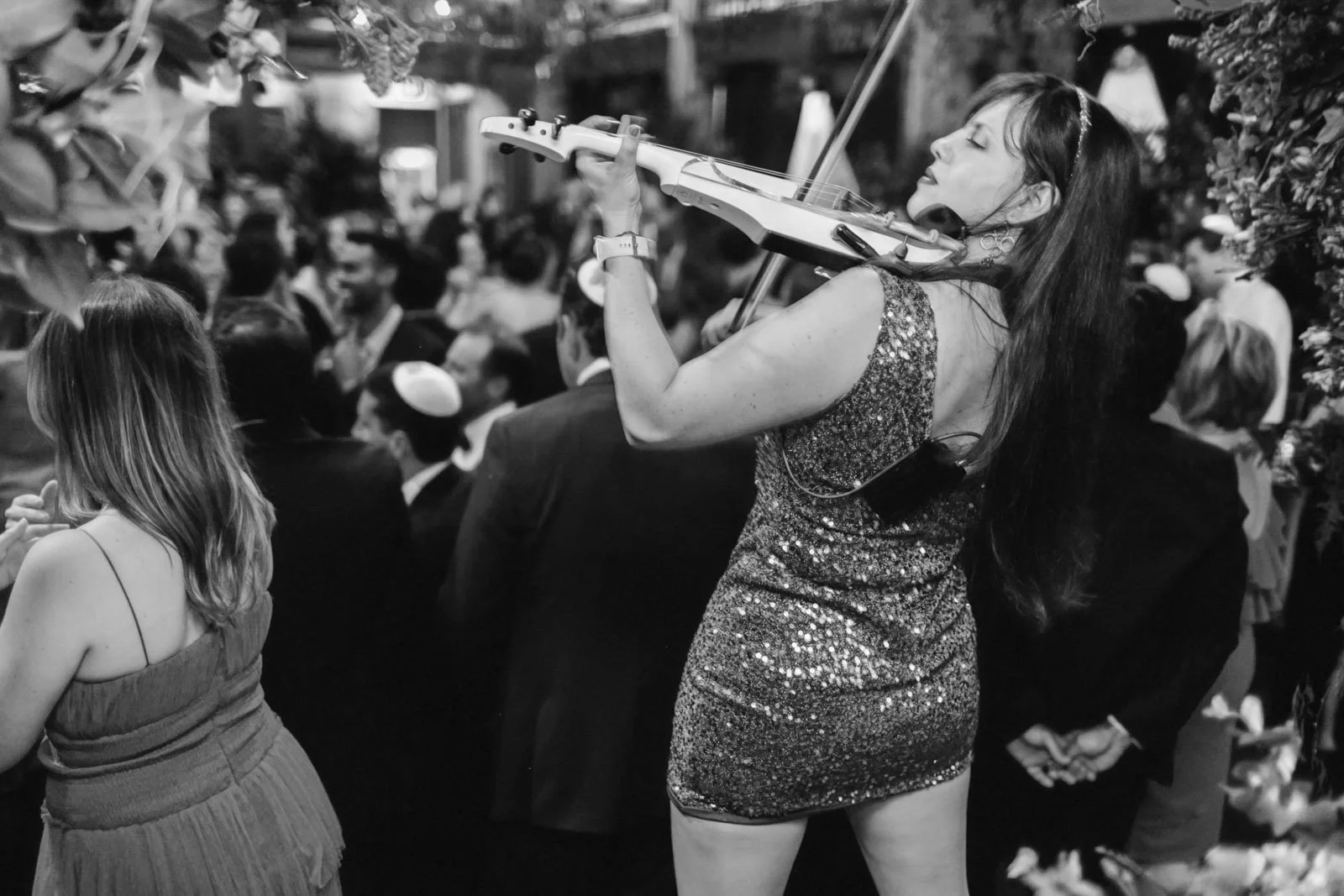 A woman in a sparkly dress plays the violin at a crowded social event, with many people in formal attire in the background.