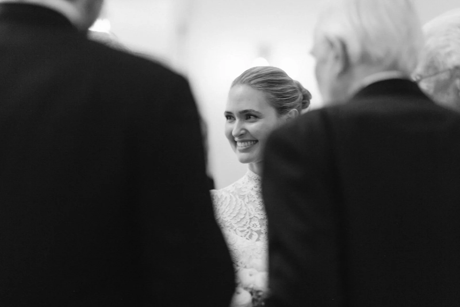 A smiling woman in a lace dress, framed by two men in dark suits, at a formal event in black and white.