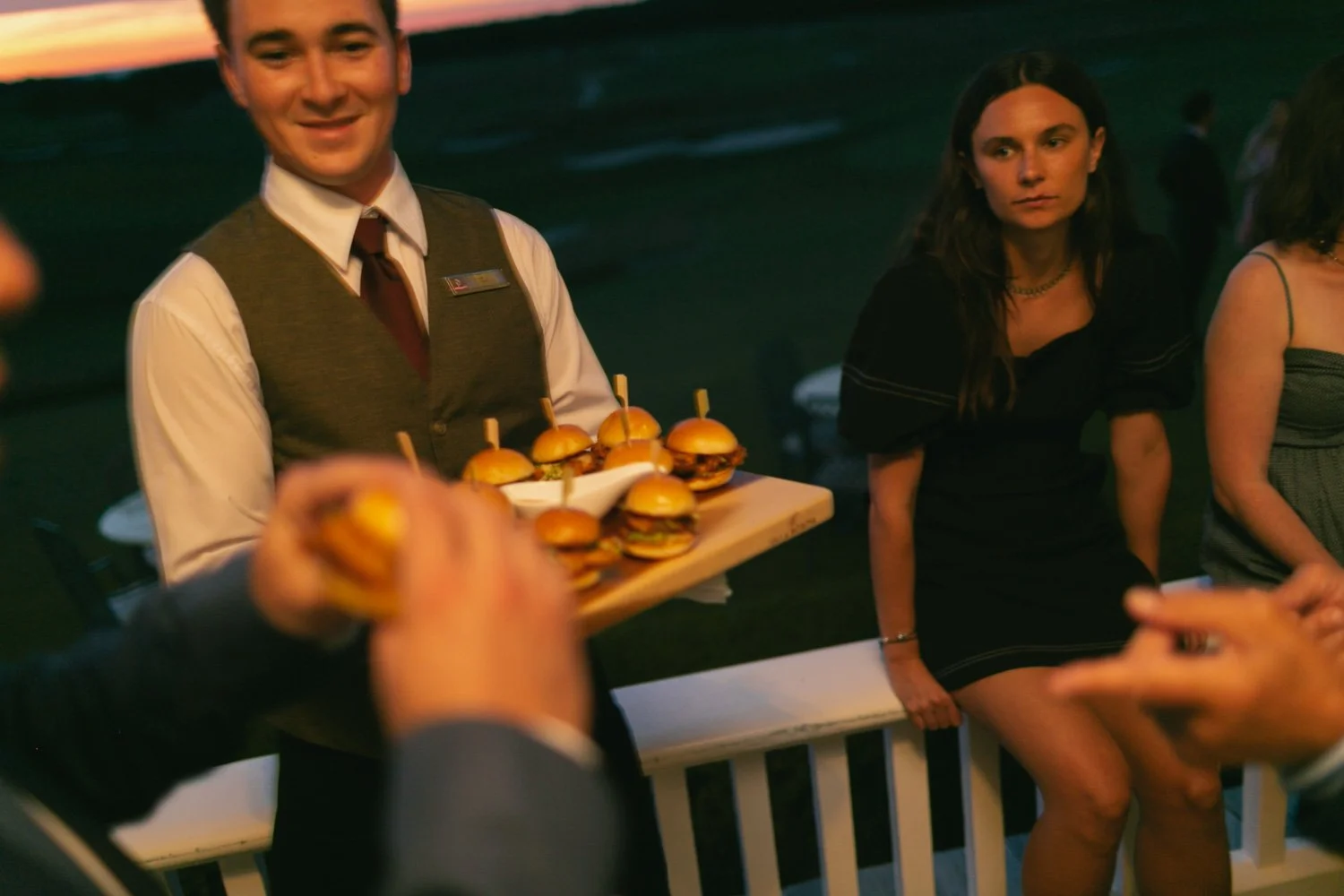 A man dressed in formal attire is holding a tray of small burgers at an outdoor evening event, while surrounded by people, including a woman in a black dress who is sitting on a white railing.