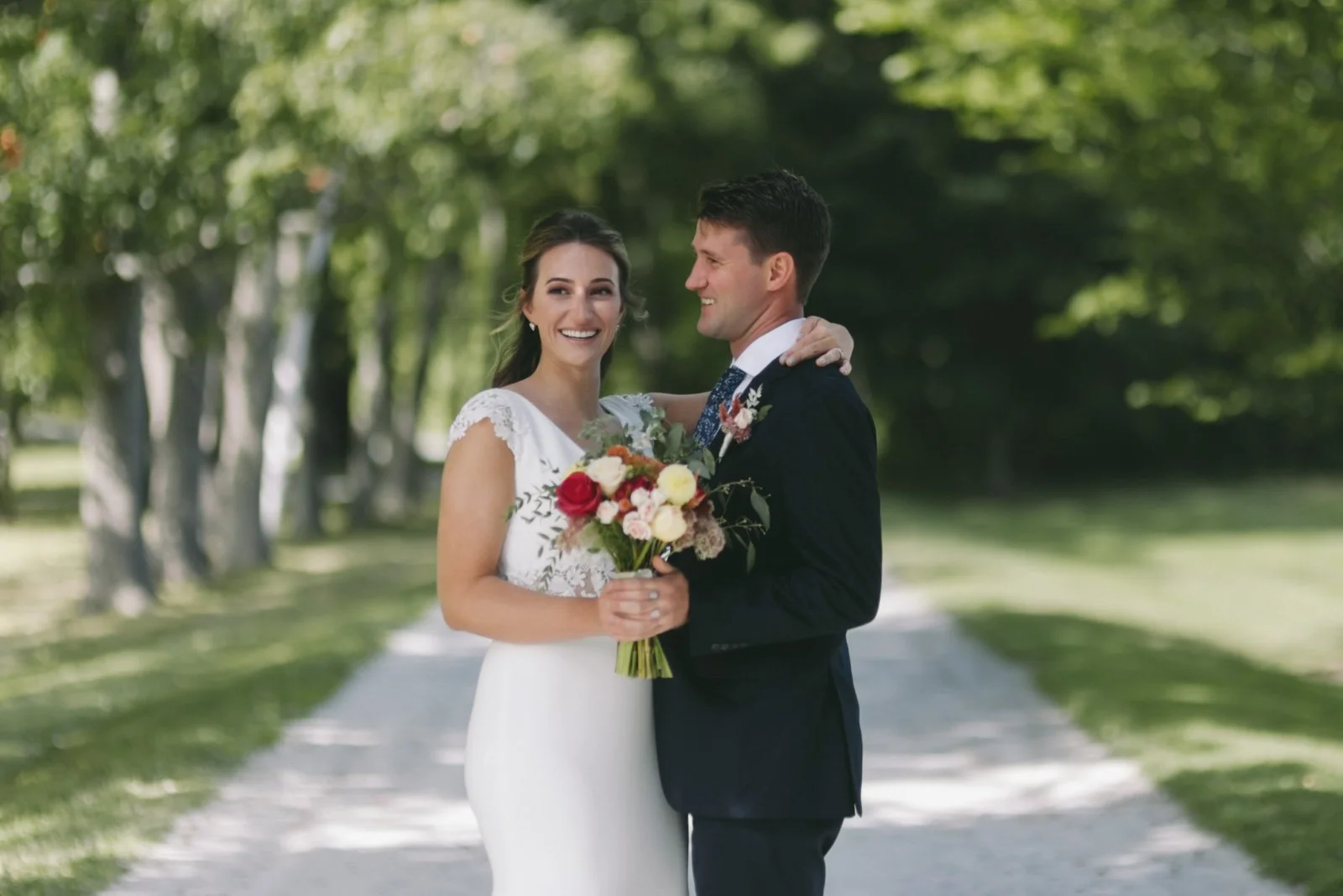 A bride and groom smiling while standing outdoors on a tree-lined path. The bride holds a bouquet of red and white flowers, and the groom has his arm around her shoulder.
