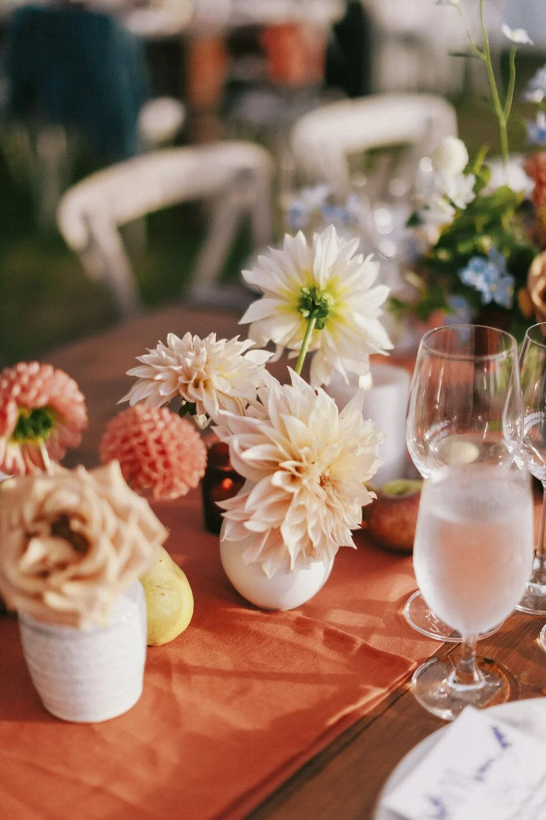 Floral centerpiece with white and peach flowers on a dining table. There are wine glasses and a table runner on a wooden table.