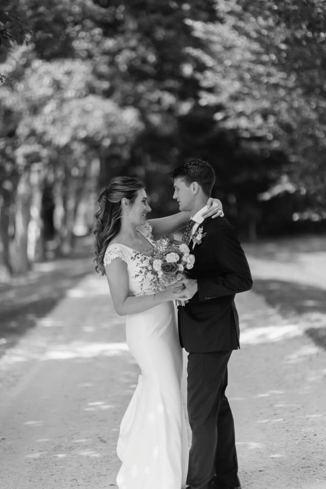 A black and white photo of a bride and groom standing face to face outdoors, smiling at each other. The bride is holding a bouquet of flowers and is wearing a long gown with lace details on the sleeves. The groom is wearing a suit and tie. They are s