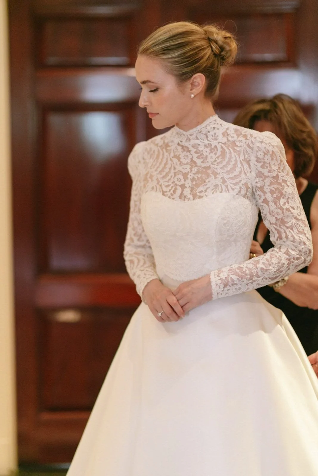 A woman in a white wedding dress with lace long sleeves and a high neckline, with her hair styled in an elegant updo, standing with her eyes closed and hands clasped.