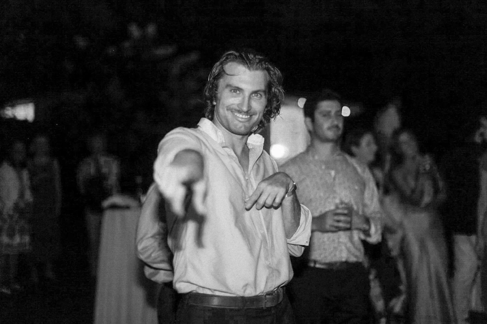Black and white photo of a man smiling and pointing towards the camera at a social event or party, with other people in the background.