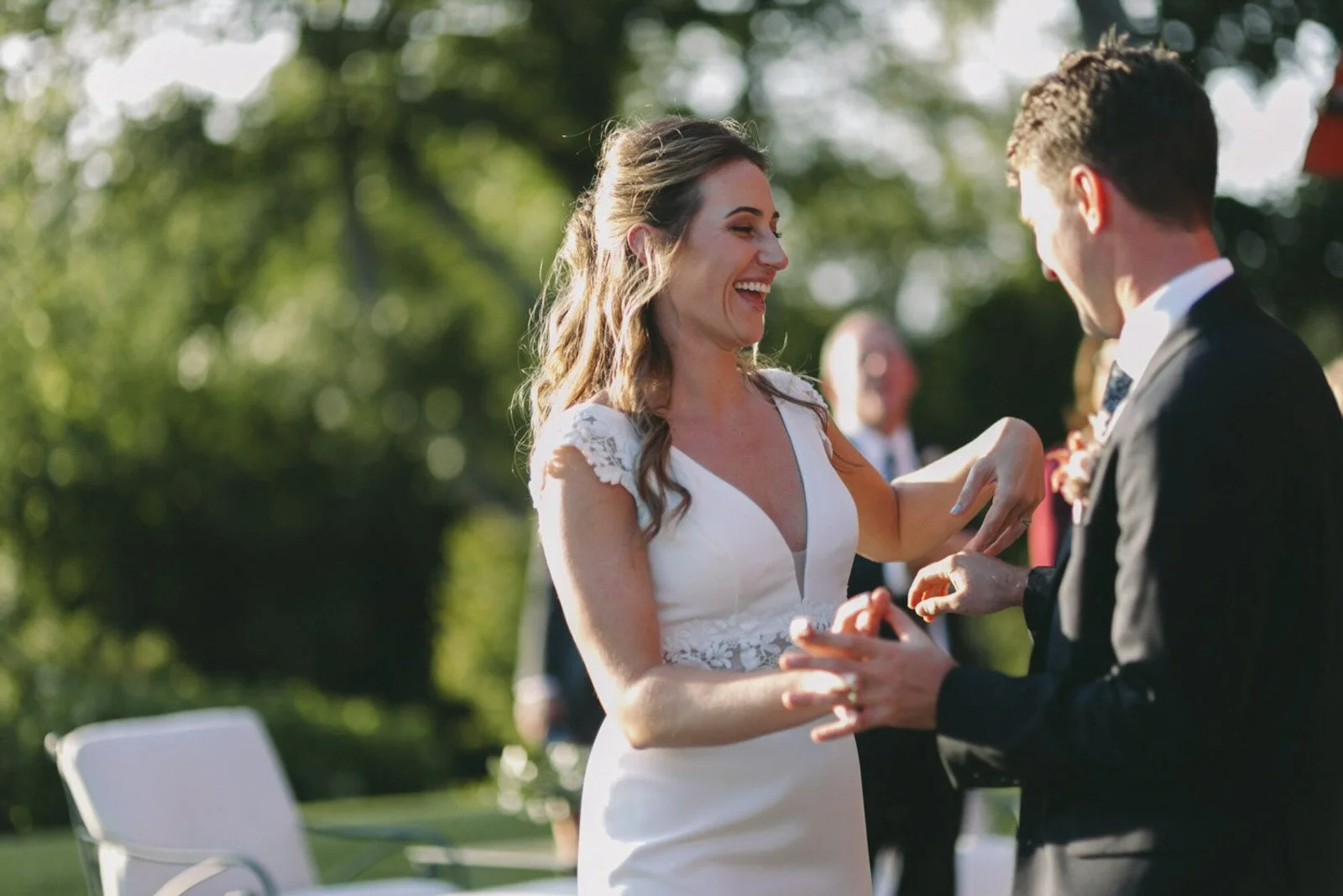 A bride and groom exchange rings outdoors during a wedding ceremony in natural sunlight, with greenery in the background.