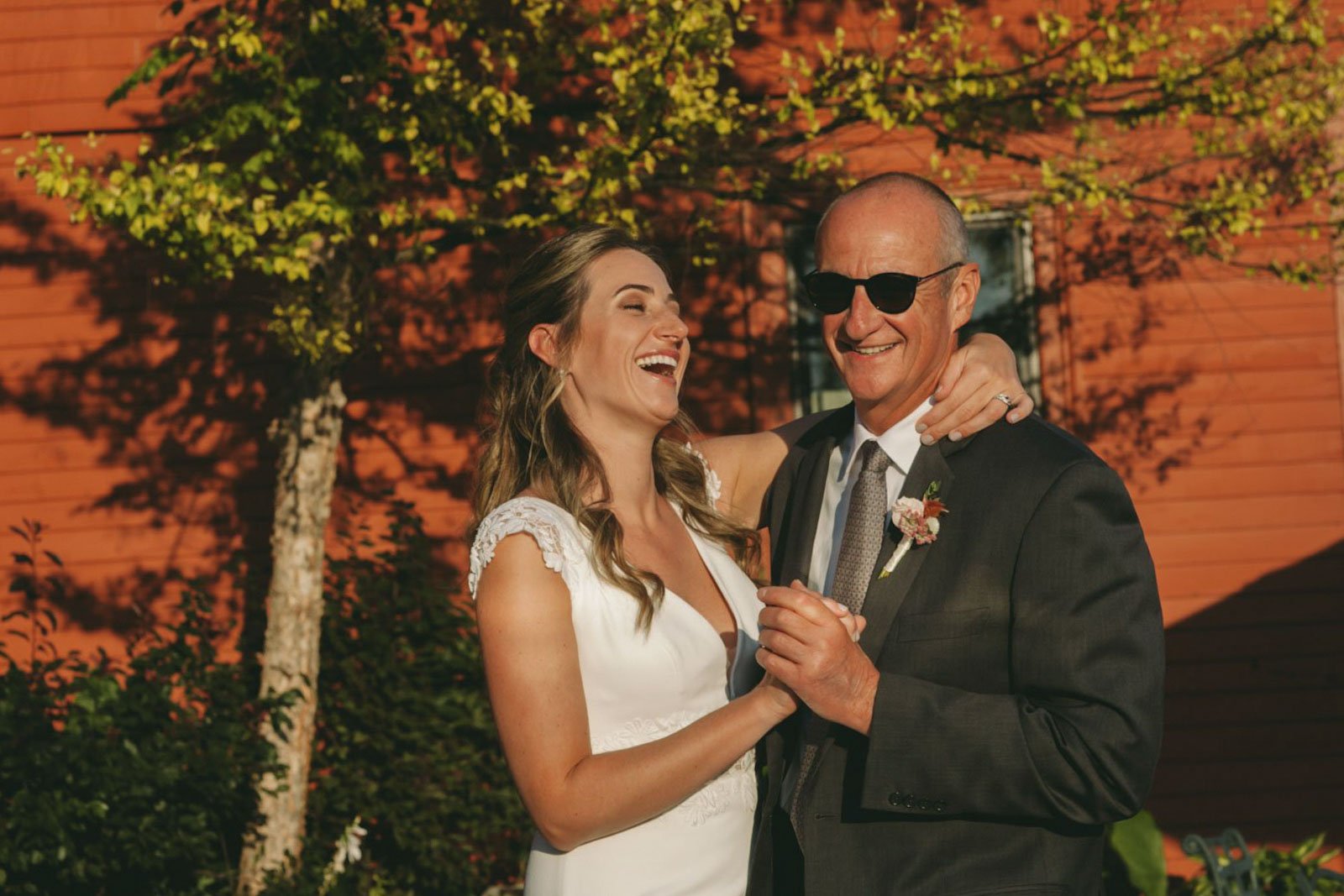 A bride and an older man, possibly her father, share a dance outdoors on a sunny day. The bride is wearing a white wedding dress, and the man is dressed in a suit with sunglasses. They are smiling and laughing, with a red wooden building and a green 