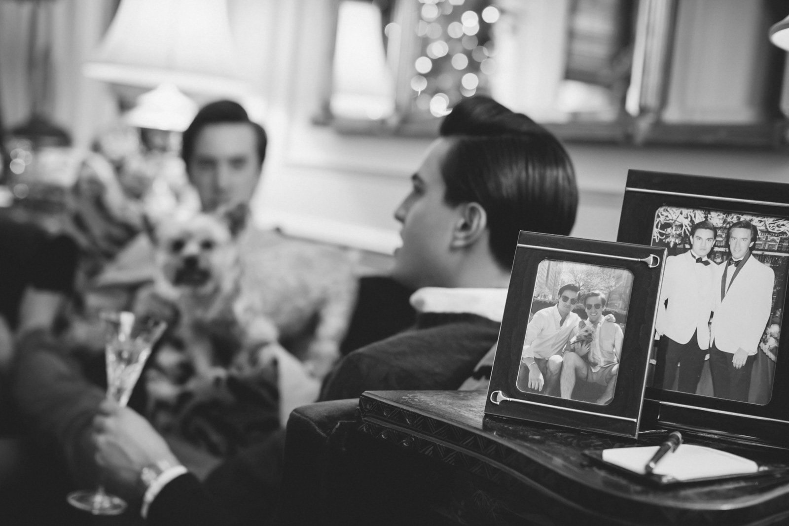 Black and white photo of a man with slicked-back hair sitting indoors. In front of him are framed photographs of two men in tuxedos and two men sitting outside, smiling. The setting appears to be a living room with windows and a decorated Christmas t