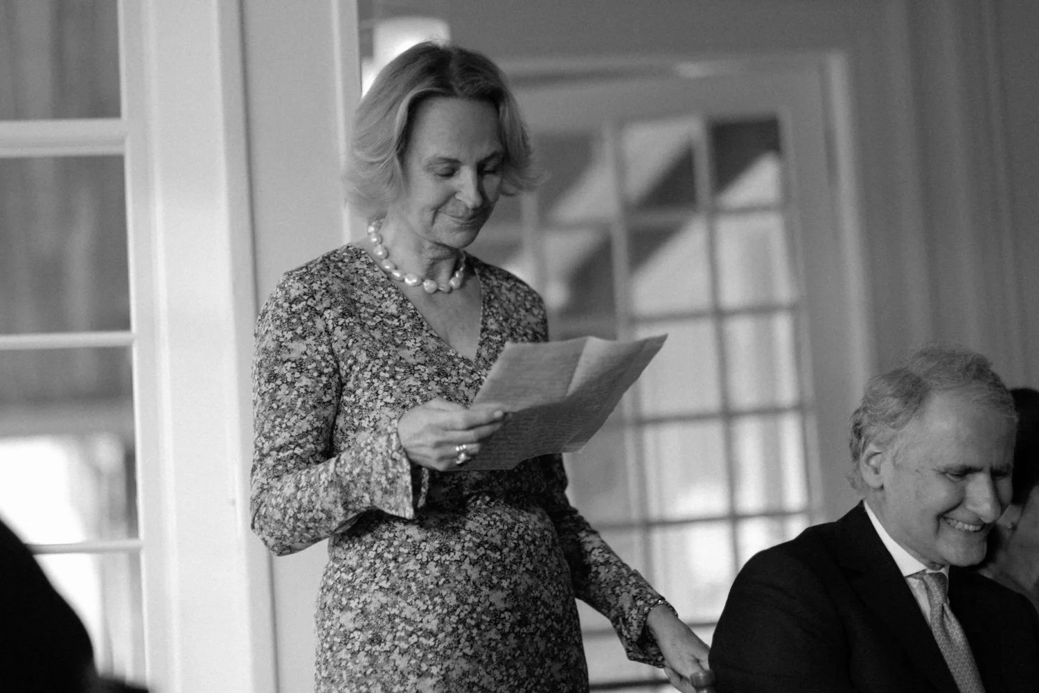 A woman in a floral dress holding a piece of paper, standing and smiling while holding hands with a man seated beside her. The scene appears to be in a room with large windows in the background.