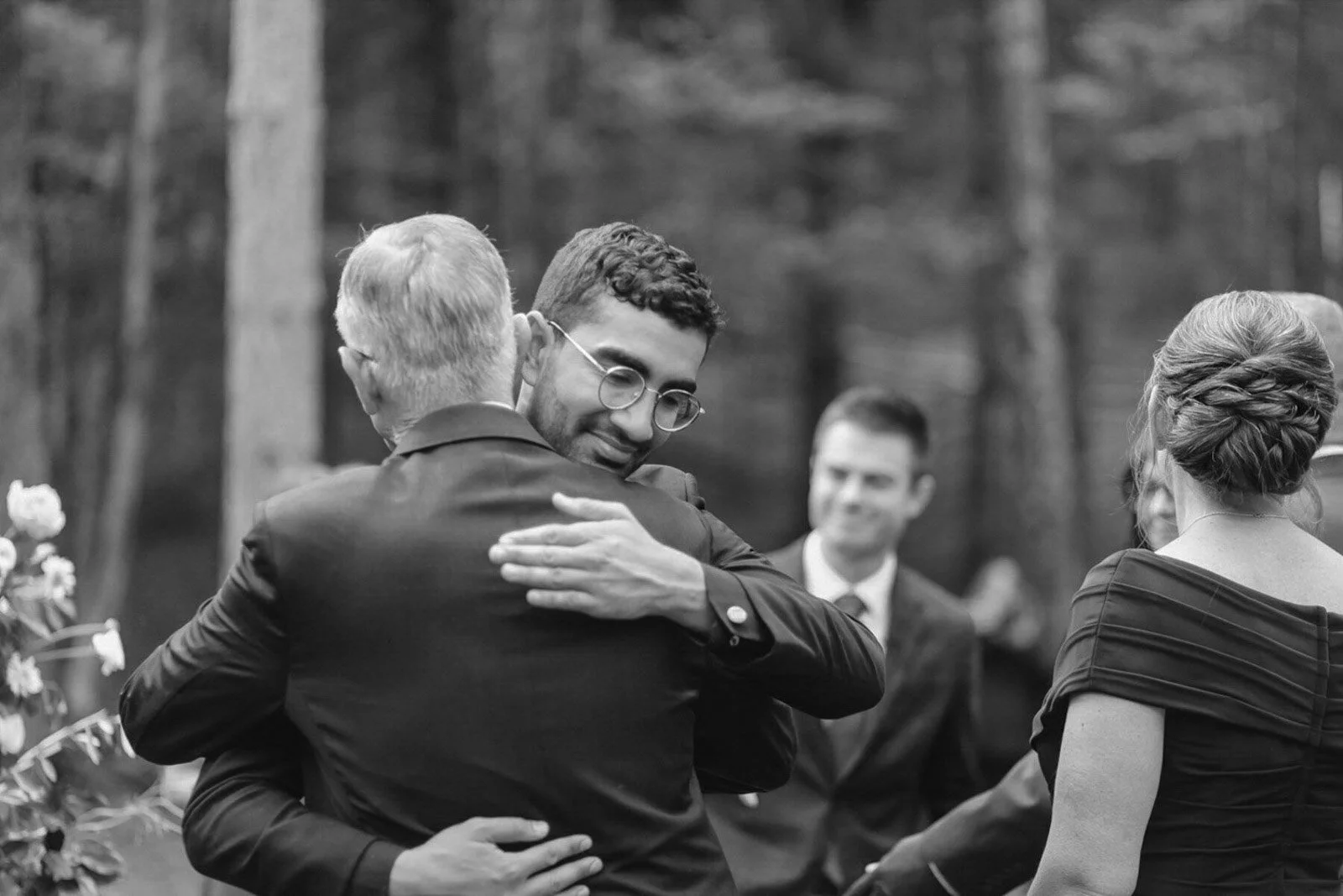 Two men hugging at a wedding ceremony, with a woman and a man in the background, outdoors among trees.