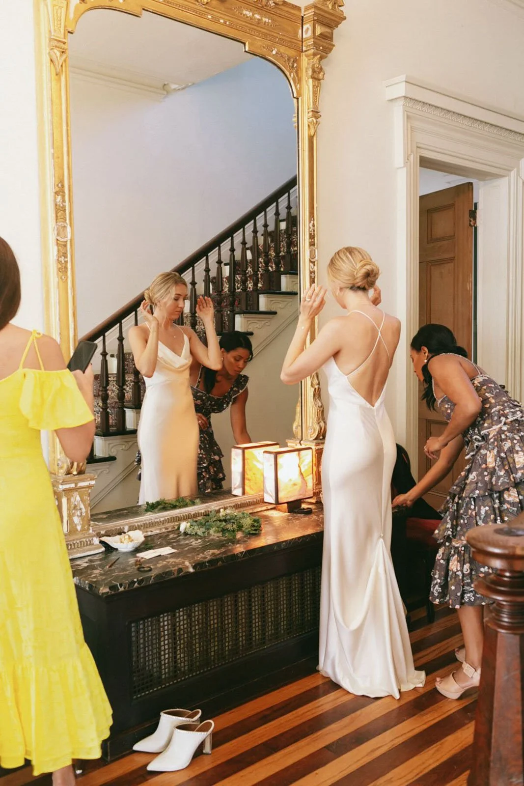 Women getting ready in a luxurious room, with a large ornate mirror, a staircase, and a marble counter with lamps, plants, and other items. Two women are dressed in white gowns, and others are preparing or fixing their dresses.