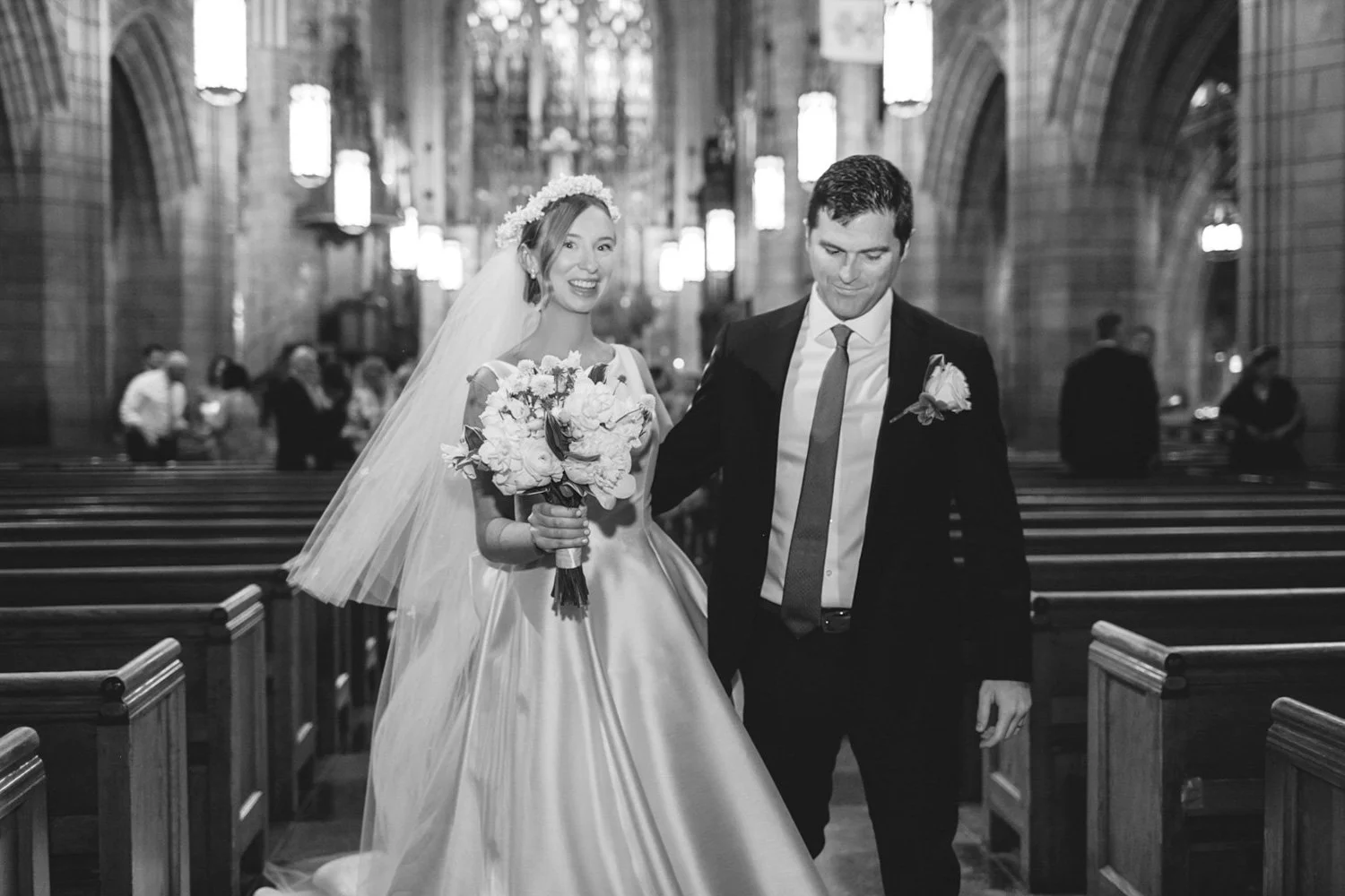 A bride and groom walking down the aisle in a church, smiling. The bride is holding a bouquet of flowers and wearing a long dress and veil. The groom is wearing a suit with a boutonniere. The church has wooden pews and stone walls.