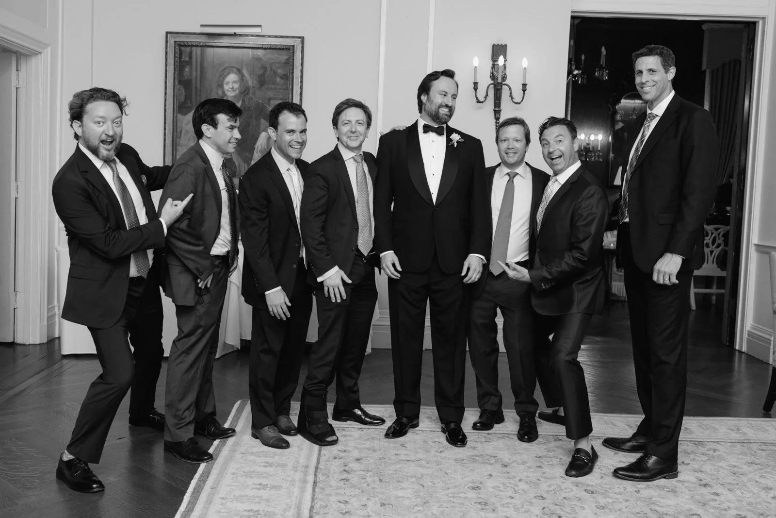 A group of men in tuxedos and suits, standing in a living room, smiling, with one man in the center dressed as a groom at a wedding.
