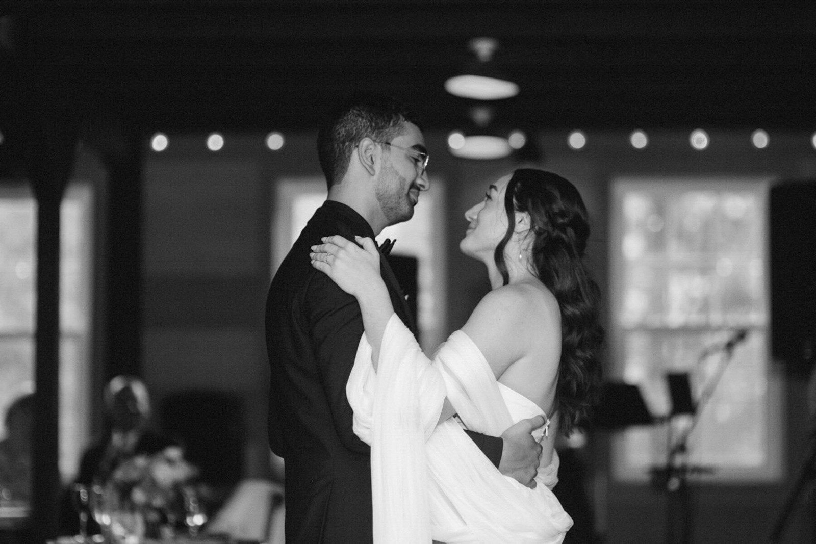 A man and woman dancing and gazing at each other at a wedding reception, with soft lighting and blurred guests in the background.