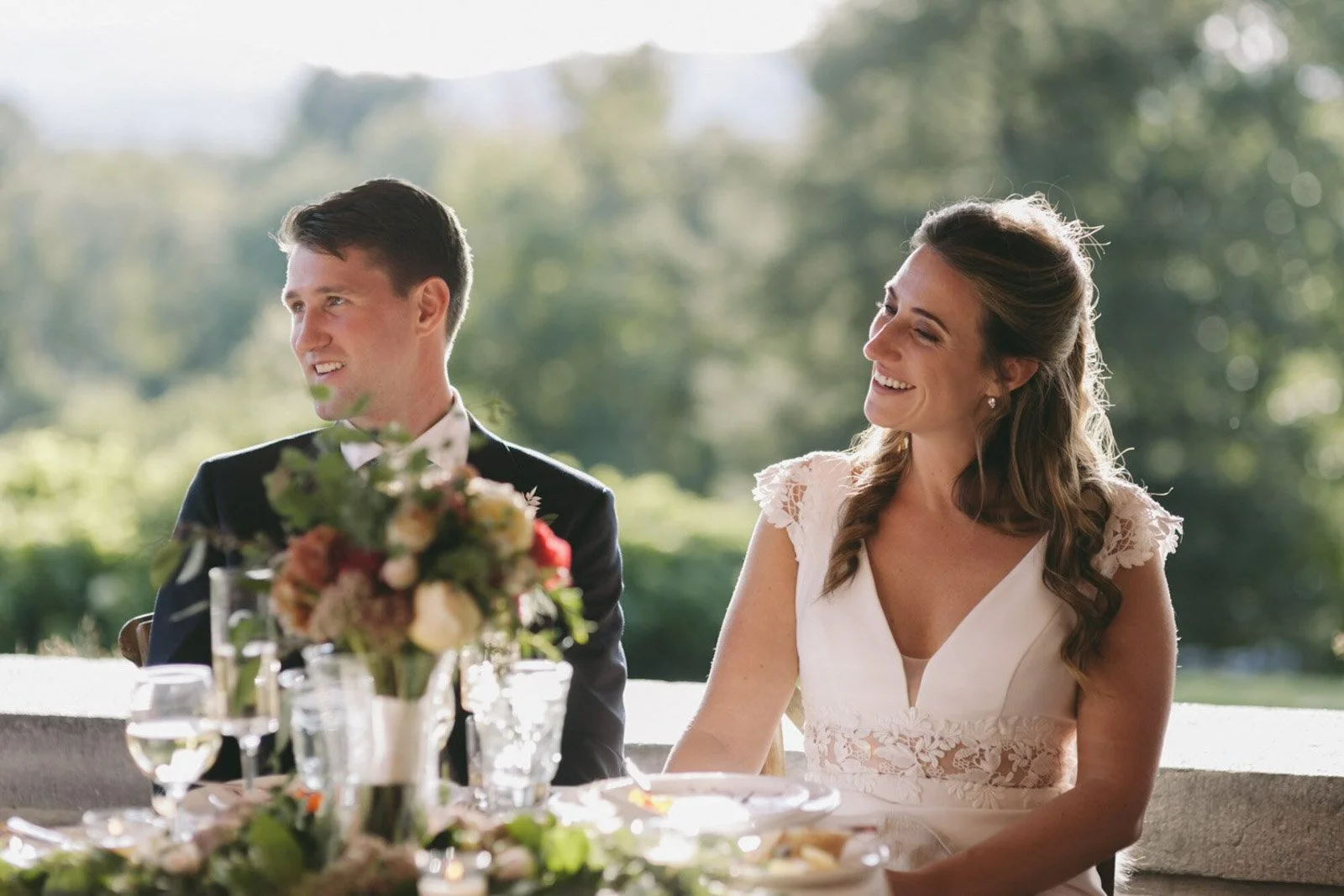 A bride and groom sitting at a table outdoors during daytime, smiling and looking towards the right, with a blurred green landscape in the background. The table has a floral centerpiece and glasses of water.