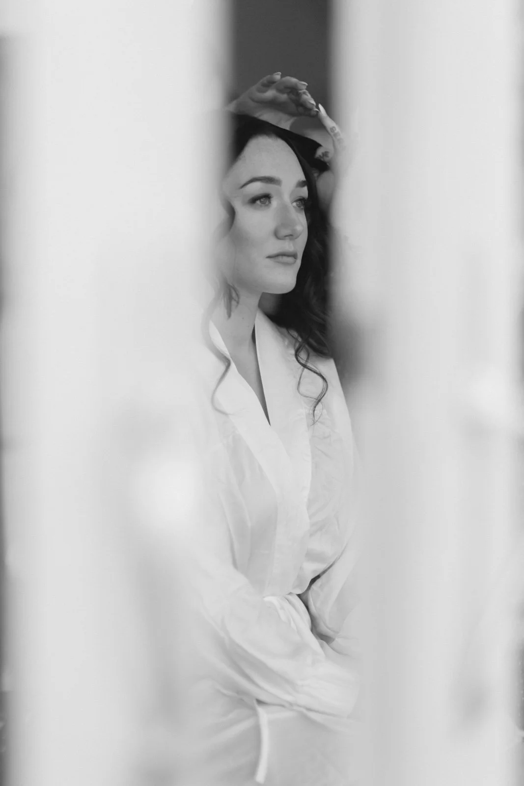 Black and white photo of a woman with dark hair, wearing a light-colored robe, looking thoughtfully through a narrow opening.