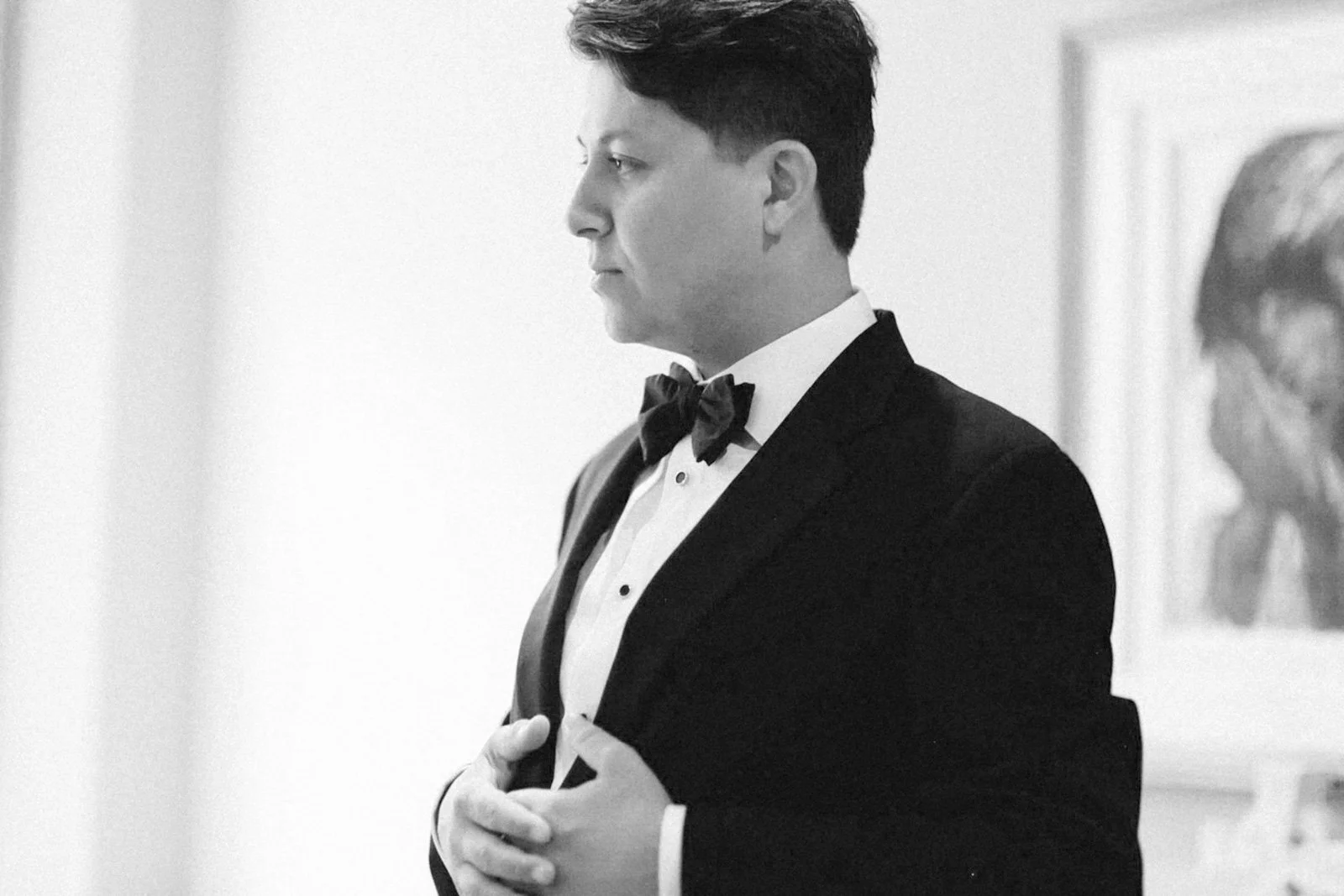 A man dressed in a tuxedo with a bow tie, standing with his eyes closed and hands placed on his chest, in a formal setting.