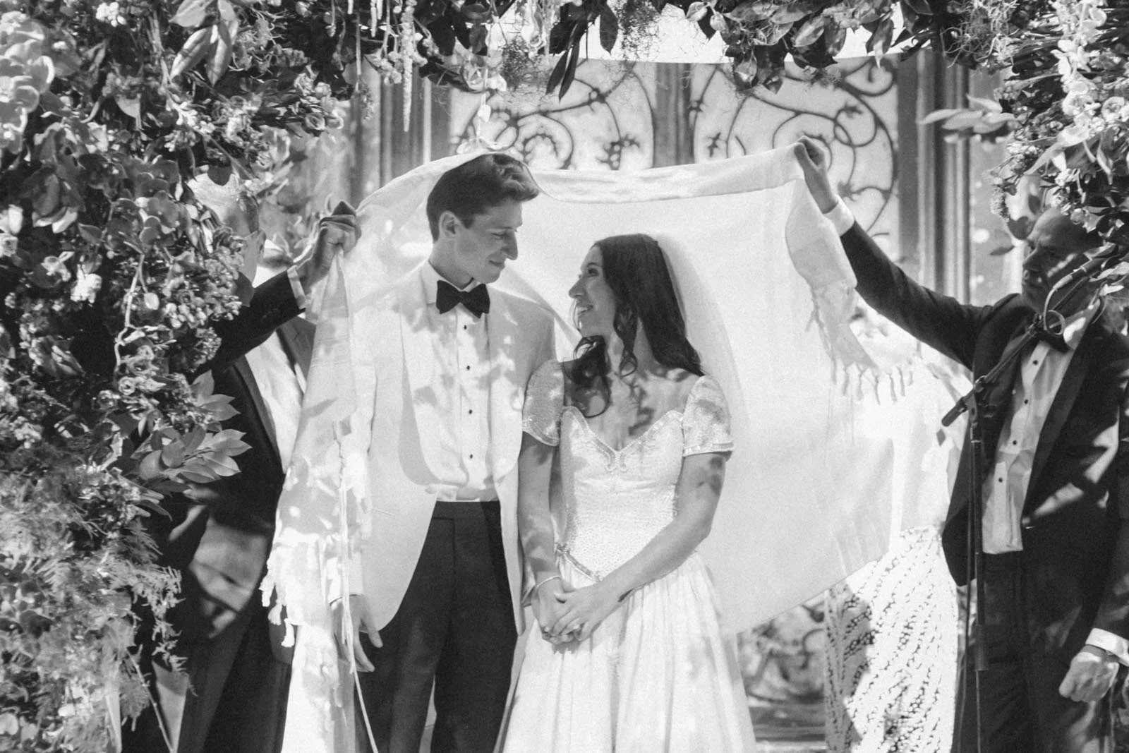 A black-and-white photo of a wedding scene with a couple facing each other under a canopy held by two men in tuxedos, surrounded by floral decorations.