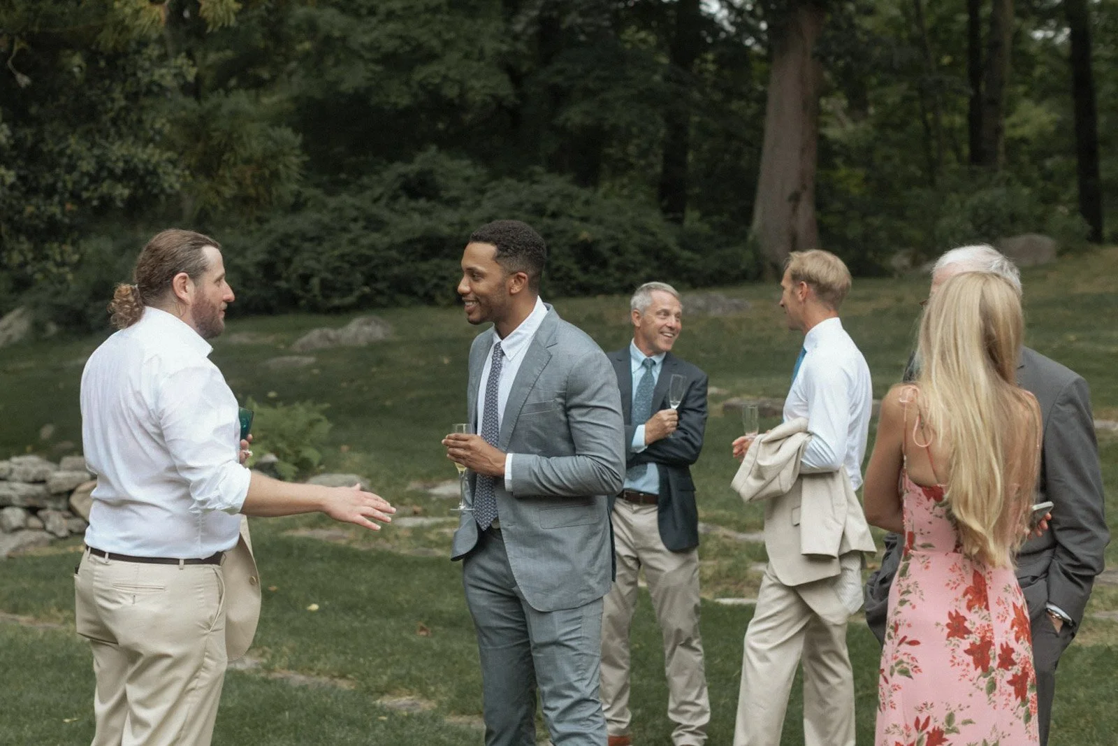 People dressed in formal wear talking and smiling outdoors in a garden setting during daytime.