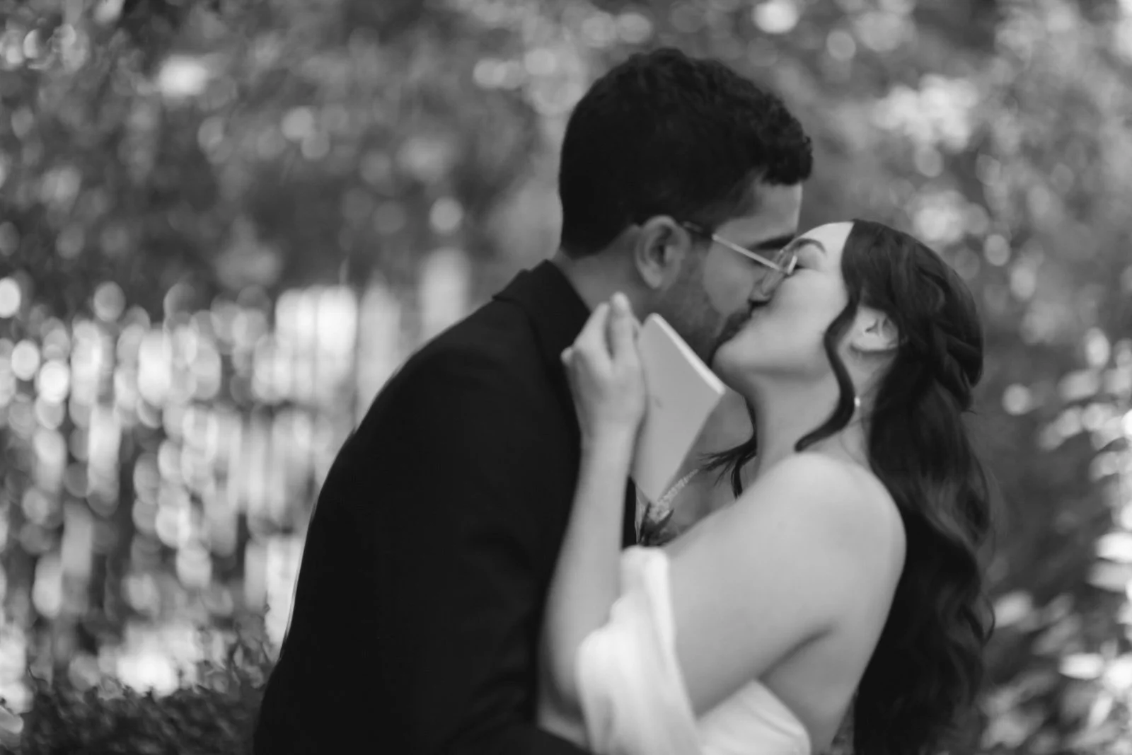 A couple sharing a kiss outdoors, with the woman in a strapless dress and the man in a suit, surrounded by blurred foliage in the background.