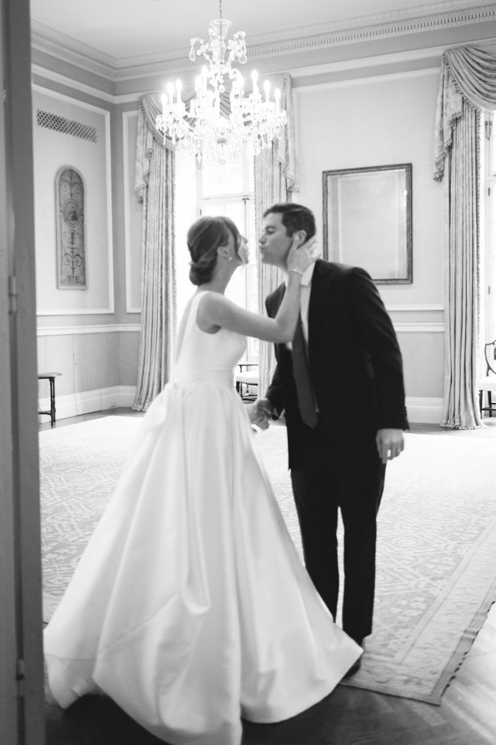 A bride and groom sharing a kiss in a luxurious, elegant room with chandelier, drapes, and ornate wall decor.