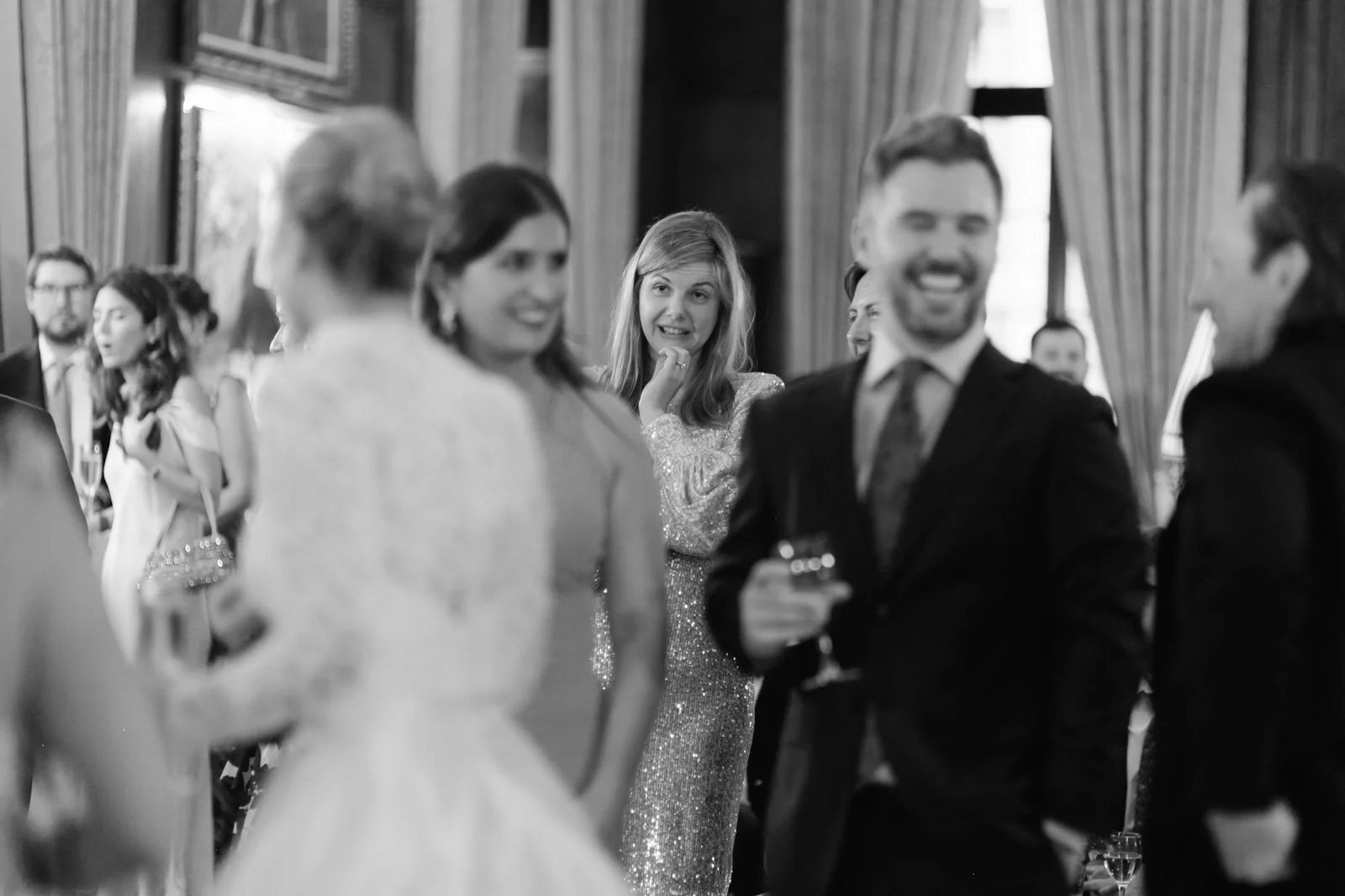 Black and white photo of a social gathering with people in formal attire, smiling and talking indoors, with curtains and windows in the background.