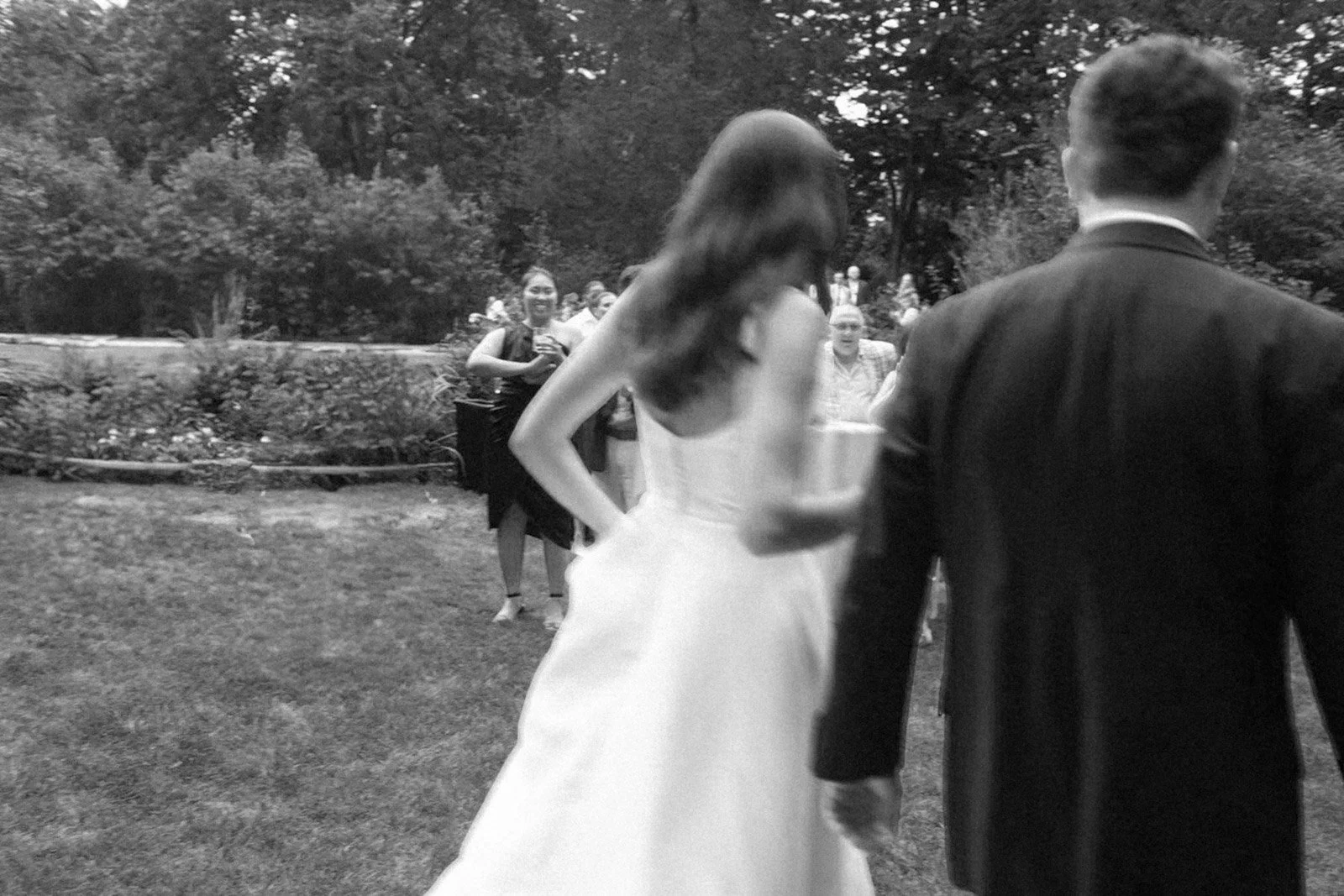 A bride and groom holding hands at an outdoor wedding reception, with guests in the background, some smiling and holding drinks.