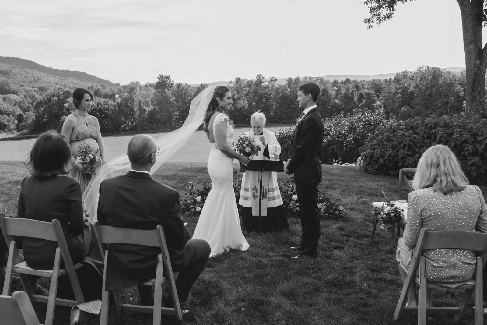 A black and white photograph of an outdoor wedding ceremony. The bride and groom stand facing each other, with the officiant reading from a book. The bride holds a bouquet, and there's a bridesmaid and a guest seated on either side. The ceremony take