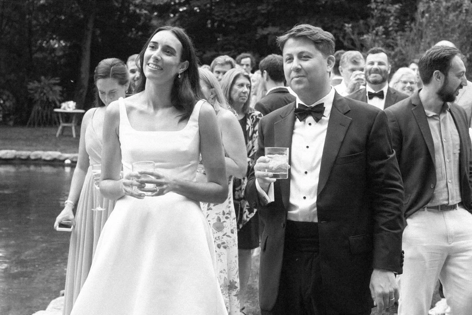 People dressed in formal attire at an outdoor gathering, some holding drinks, with a woman in a white dress and a man in a tuxedo in the foreground.