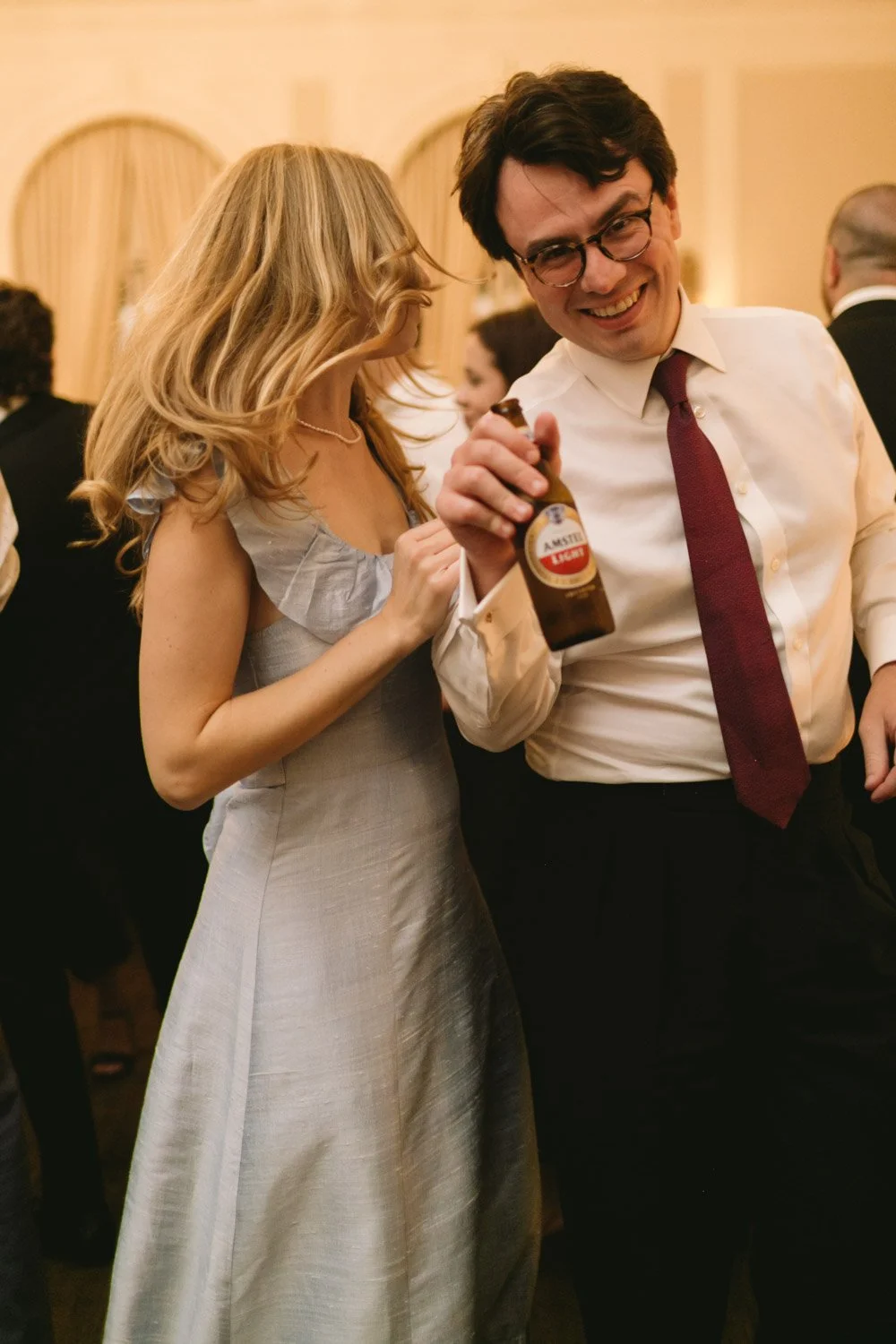 A man and woman at a social event, smiling and holding a beer bottle, with other guests in the background.
