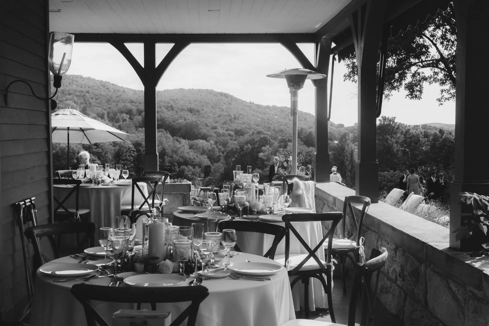 Black and white photo of an outdoor dining area with tables set for a meal, overlooking a scenic mountainous landscape with trees. The tables have glasses, plates, and utensils, and some have tablecloths. There is a large patio heater and a parasol i