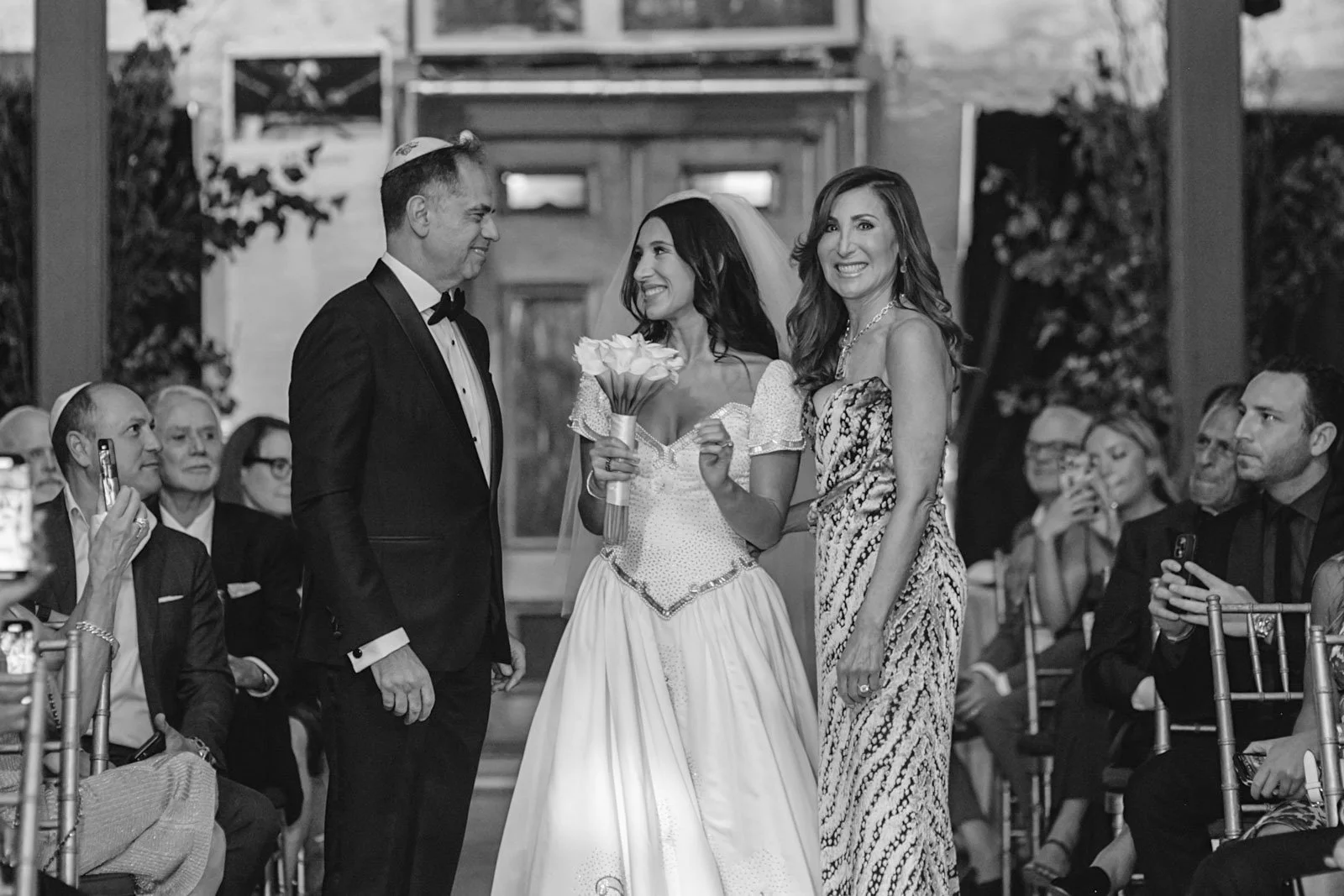 A wedding ceremony with the bride, groom, and a woman standing together, surrounded by seated and standing guests, some taking photos, in an indoor venue.