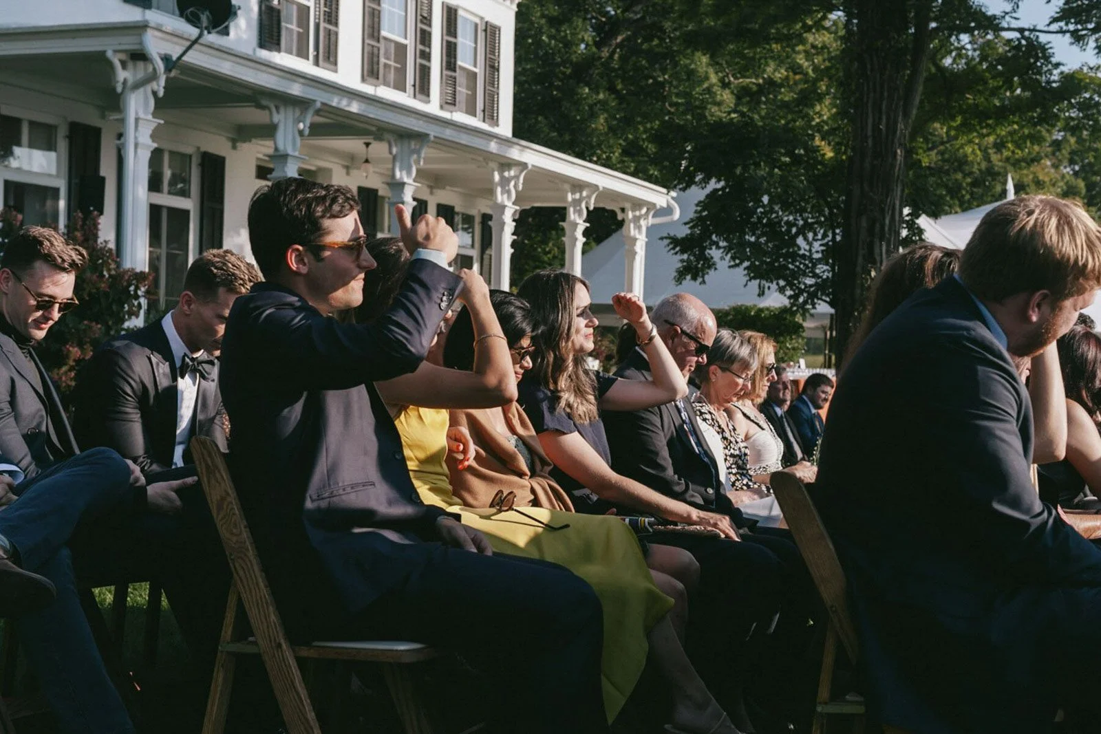 People attending an outdoor event seated on chairs in front of a white house with columns, some engaging with each other and others looking forward, in the late afternoon sunlight.